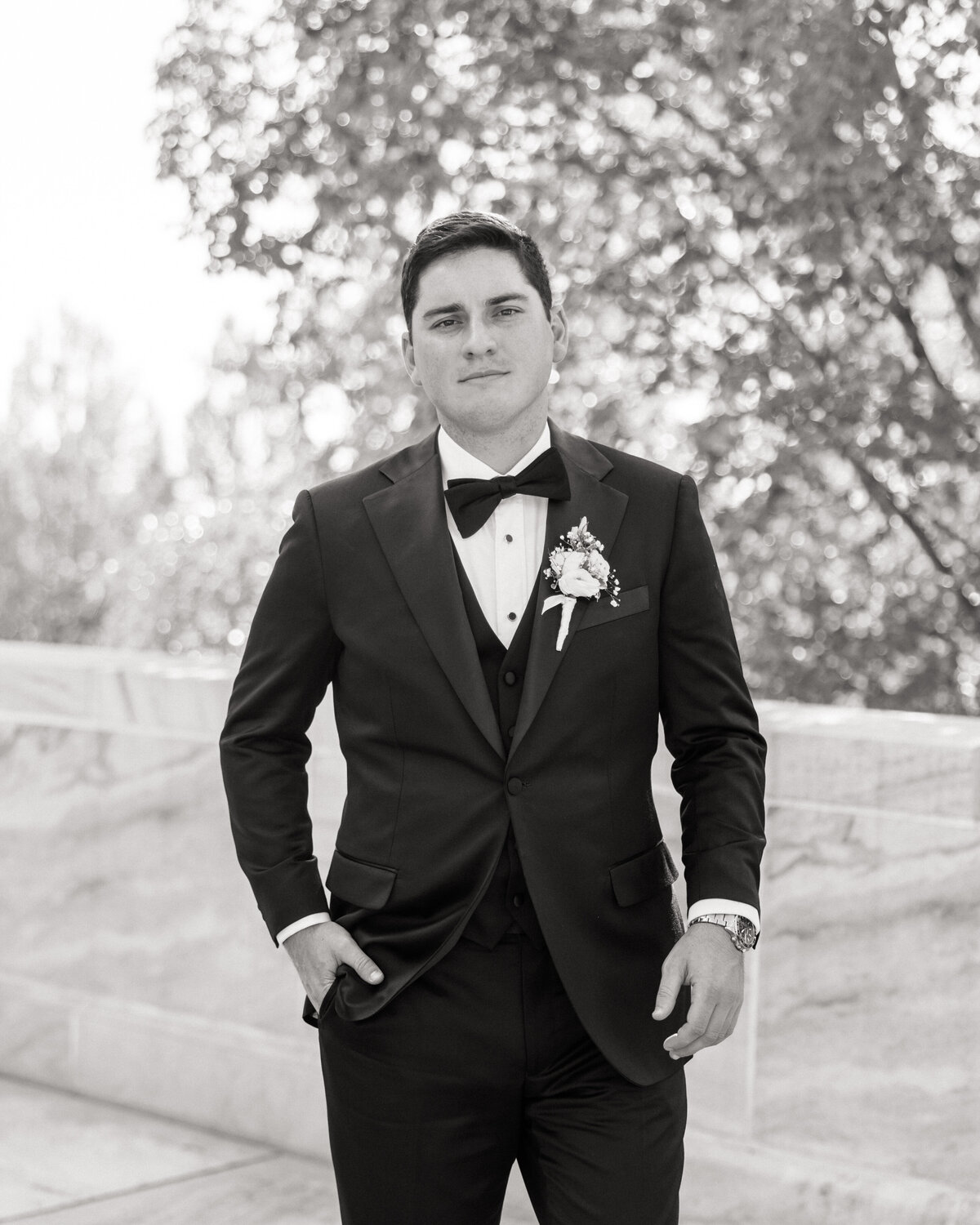 Groom poses confidently in a classic black tuxedo, boutonnière pinned to his lapel, captured by a Philadelphia wedding photographer in soft black and white.