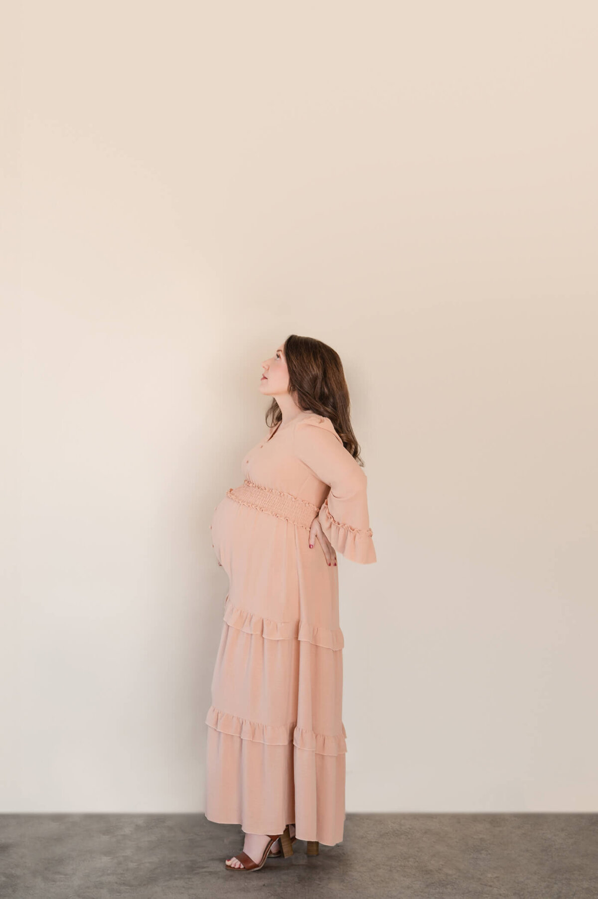 woman looking up at the sky while holding her pregnant belly getting professional maternity photos taken in a tan dress