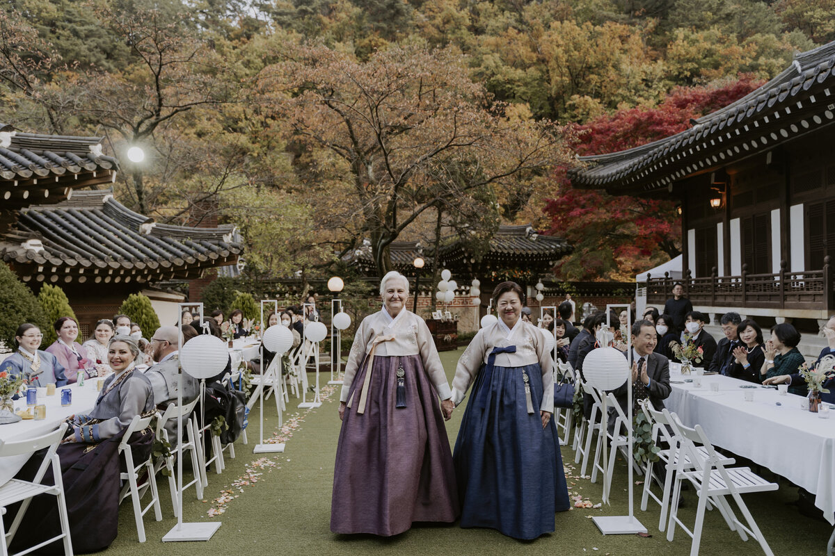 mothers in purple and blue hanbok walk hand in hand down the aisle