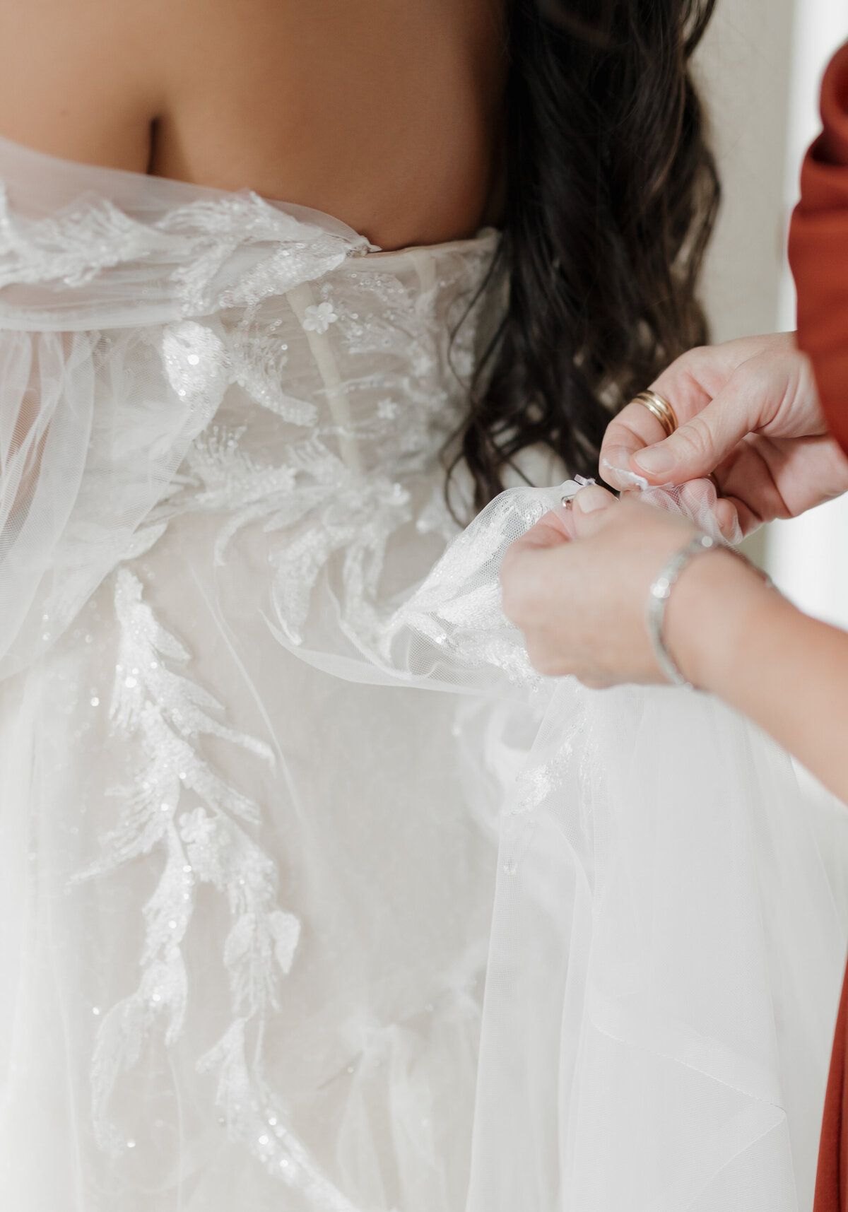 Close-up of a bride in a white wedding dress with intricate lace details. Captured by an expert in New England photography weddings, a person in a red outfit, wearing a bracelet, adjusts the fabric as the bride's long, dark hair cascades down her back.