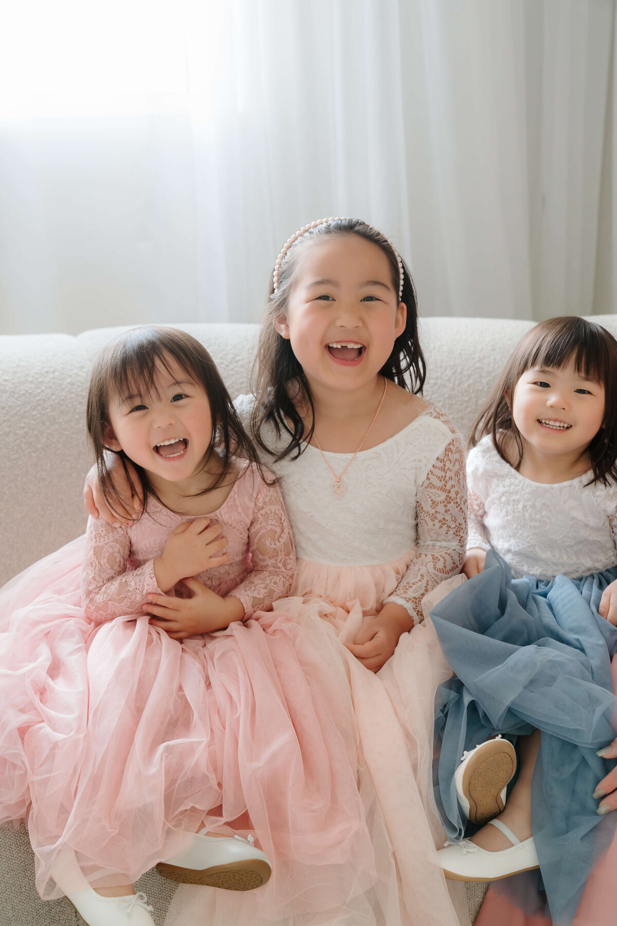 Three little girls in elegant dresses sit together on a plush couch, sharing smiles and laughter in a cozy setting.
