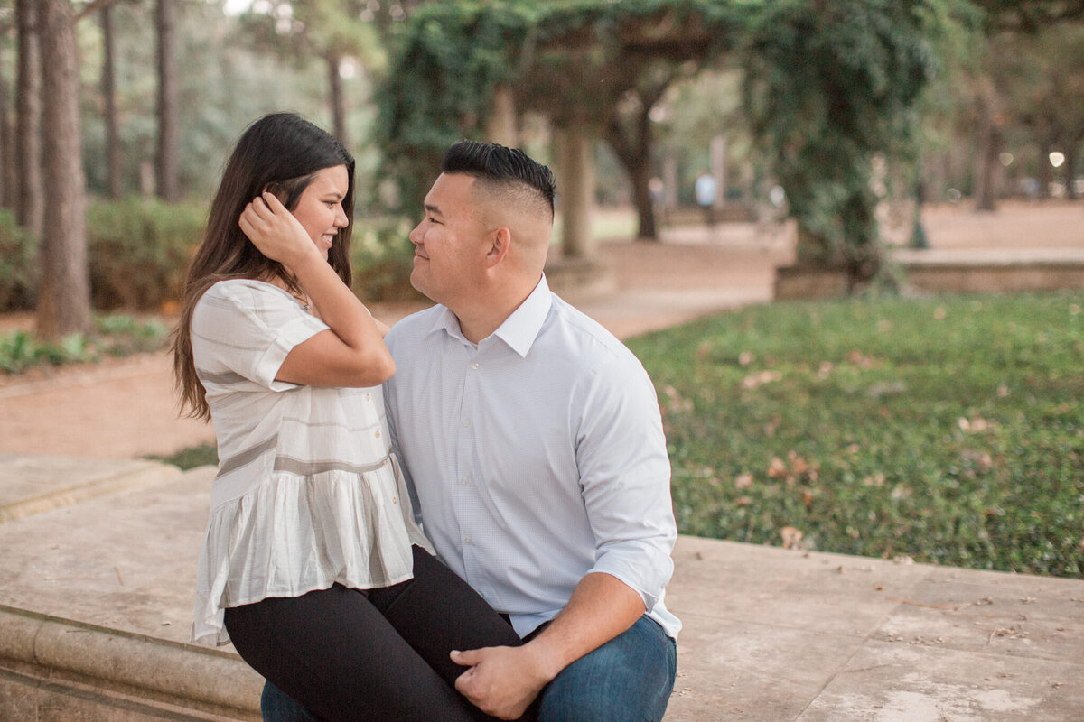 An engaged woman sits on her fiancé's knee, while tucking her hair behind her ear