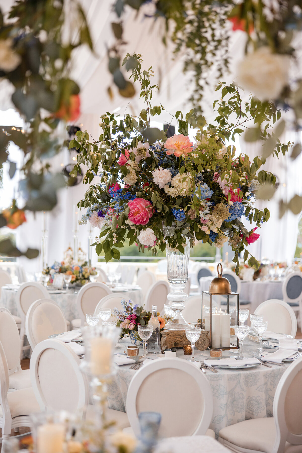 Reception tables at Water Works Philadelphia set with white chairs, crystal glassware, and lush floral centerpieces in pink, blue, and ivory tones, with greenery suspended from the tent ceiling to create a romantic garden-inspired atmosphere.