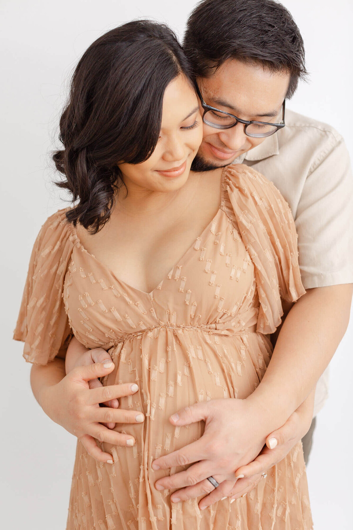 Soon-to-be Dad is standing behind Pregnant woman and wrapping arms around her belly. Their heads are touching at the foreheads and looking down at her belly with their hands on top of each others. They both have soft grins on their faces.