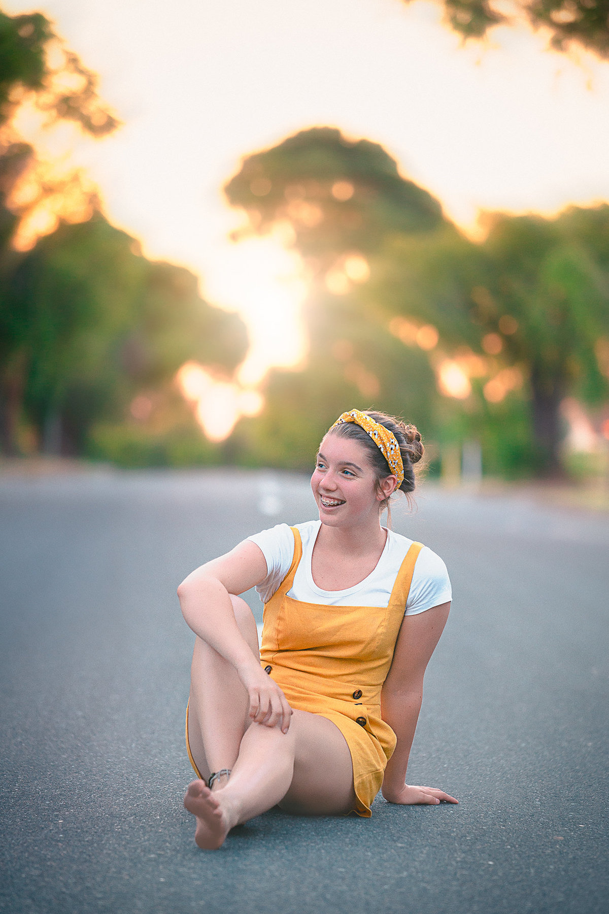 Senior picture photographers near me captures girl and a white shirt and yellow overalls sitting on a road and smiling while looking off to the distance for her senior pictures