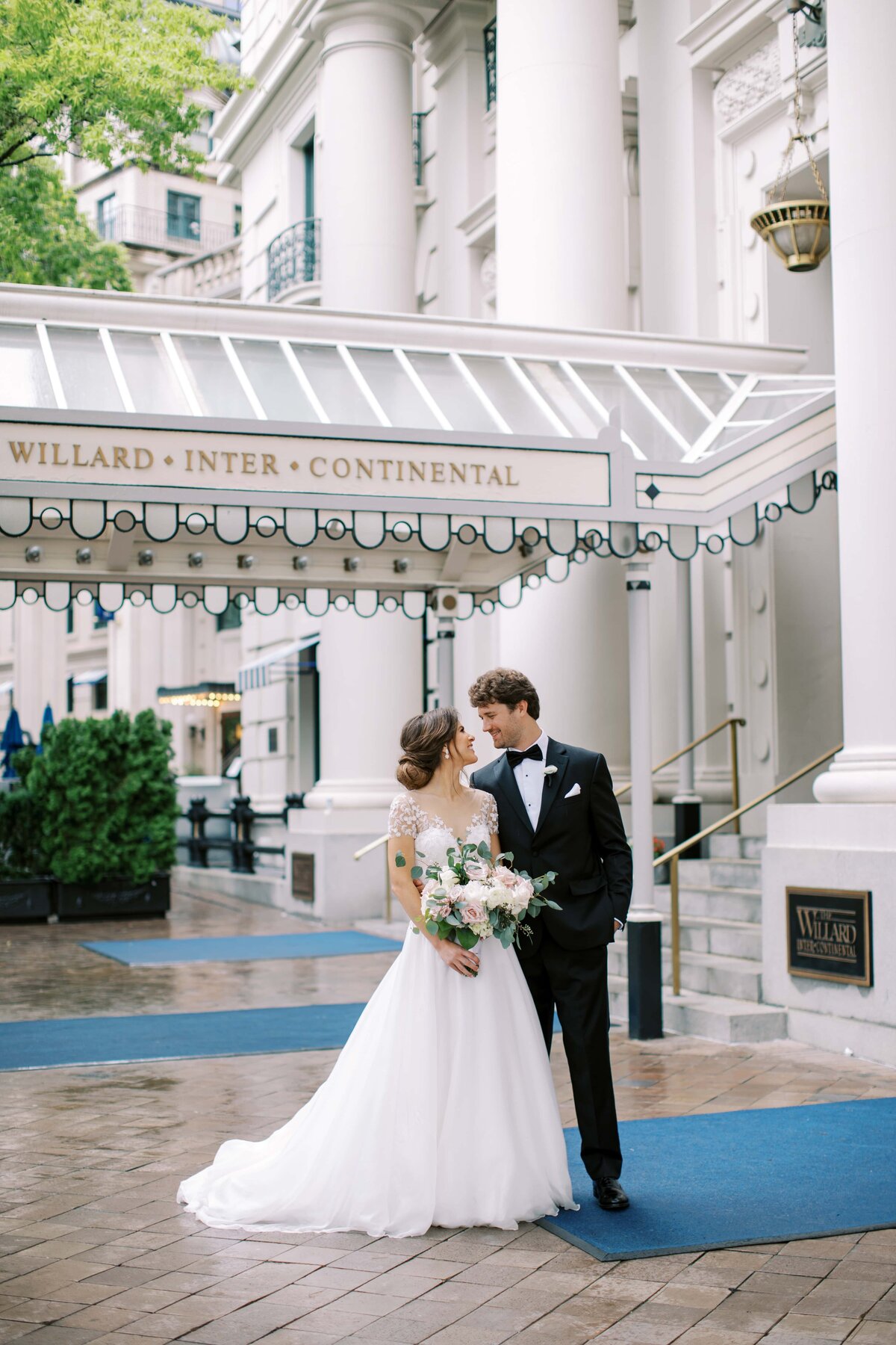 Young newlywed couple in wedding attire smiling at each other while standing side by side in front of The Willard Hotel in Washington DC