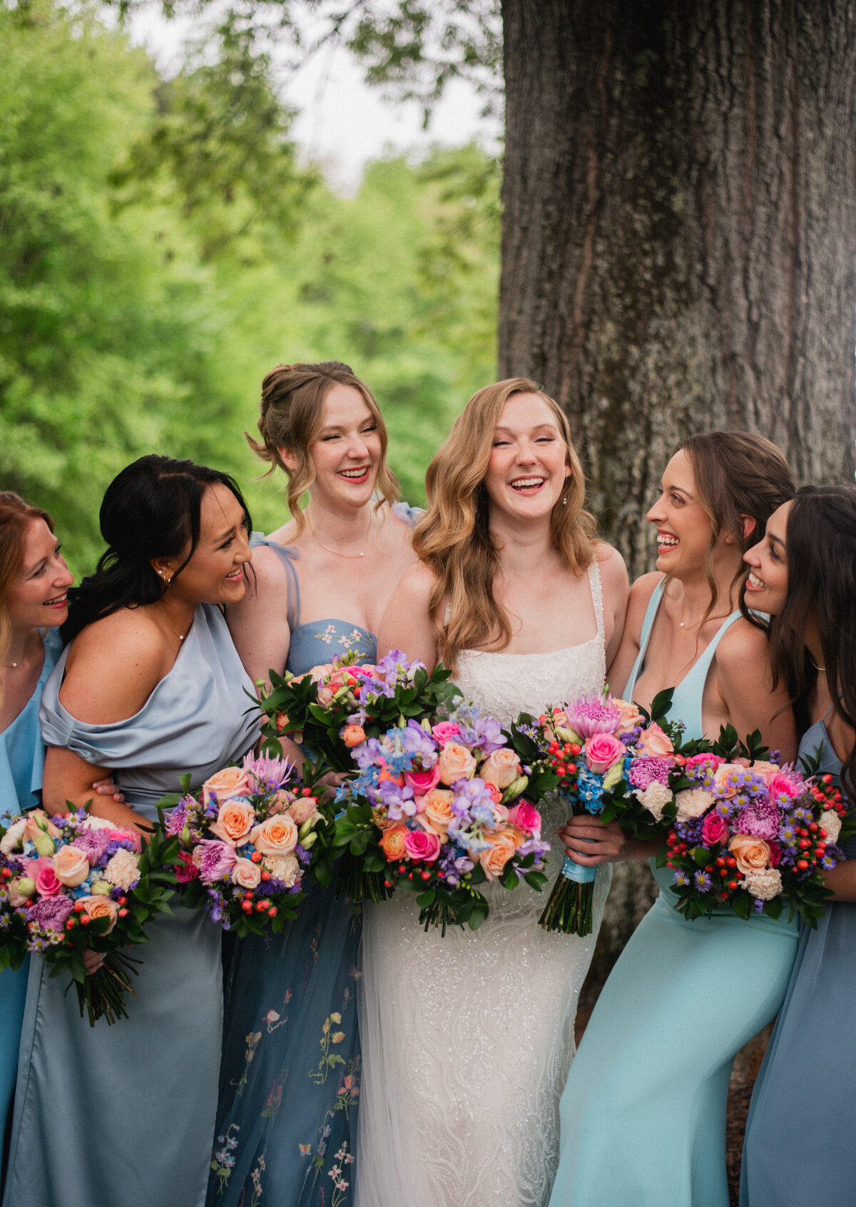 Five bridesmaids wearing dresses of different shades of blue surround a bride wearing a white dress standing in front of a tree