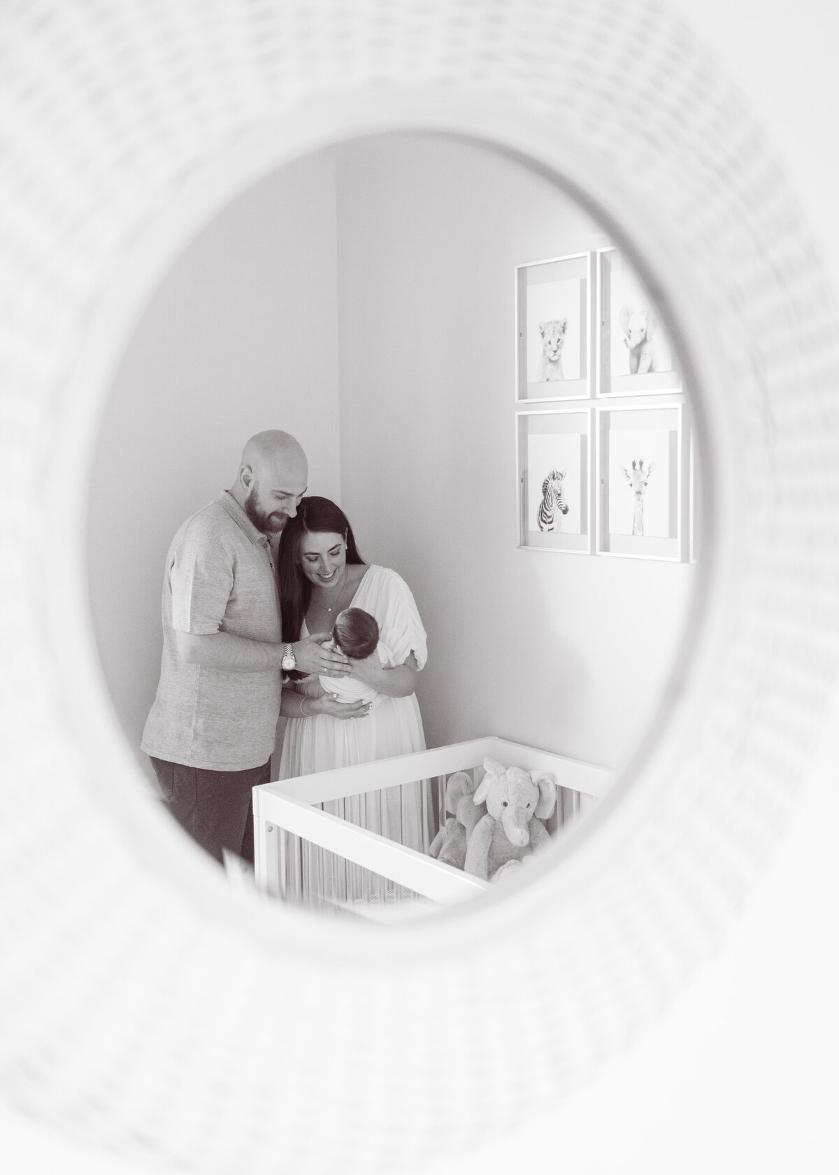 Black and white portrait of a man and woman reflected on a round mirror holding a newborn baby next to a crib.