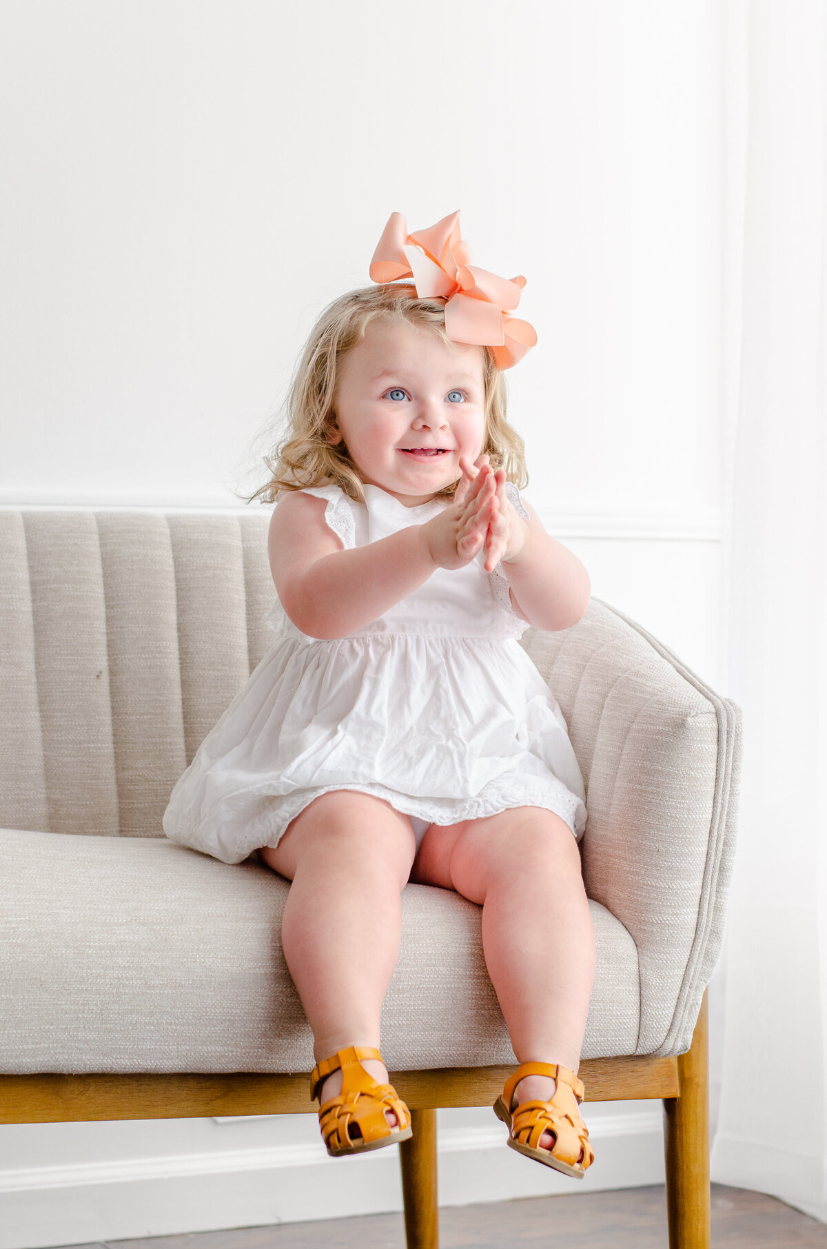 toddler claps her hands sitting on a couch in a white studio