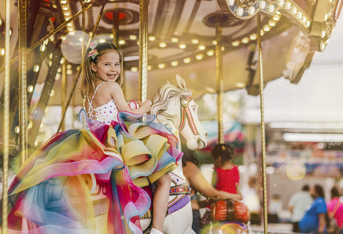 Girl in pigtails wearing a rainbow dress on a carousel horse at a carnival and smiling near Annapolis Maryland