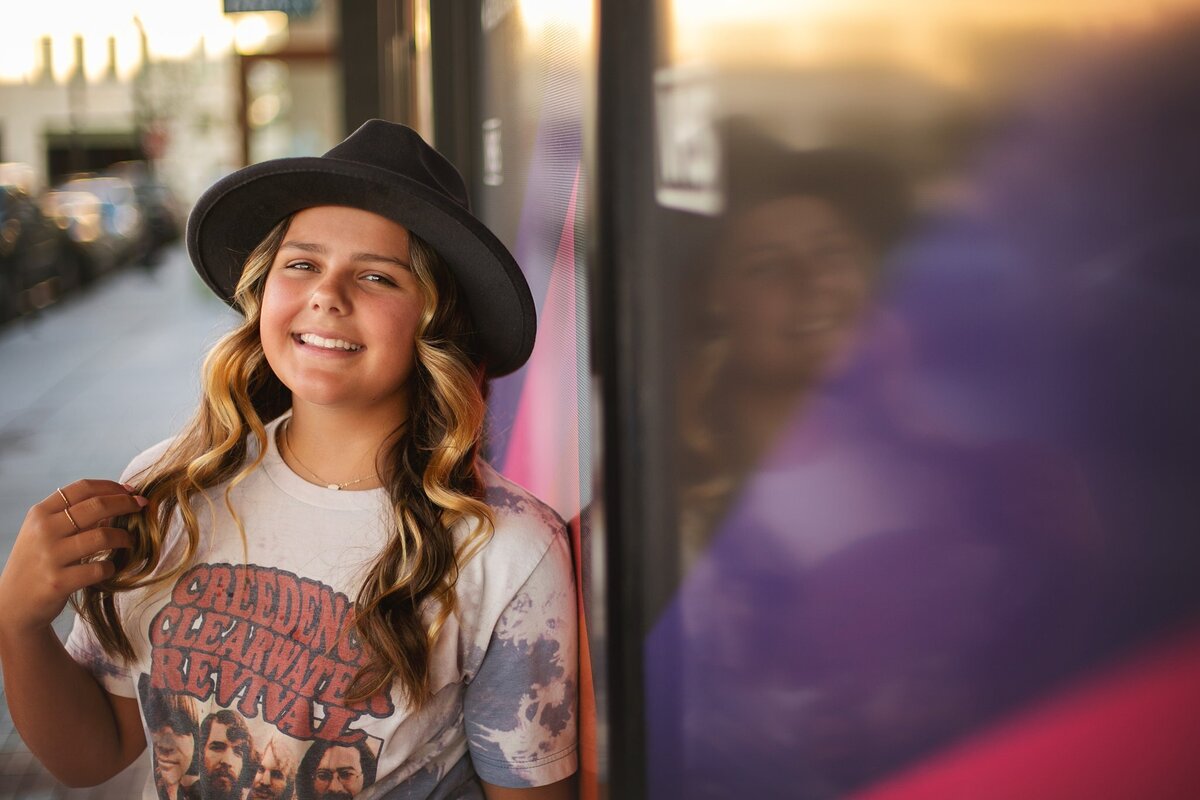 Trendy-teenage-girl-wearing-a-black-hat-and-a-vintage-band-t-shirt-smiling-in-the-warm-golden-hour-light-in-downtown-indianapolis-indiana-bottleworks-area-during-a-high-school-senior-portrait-session