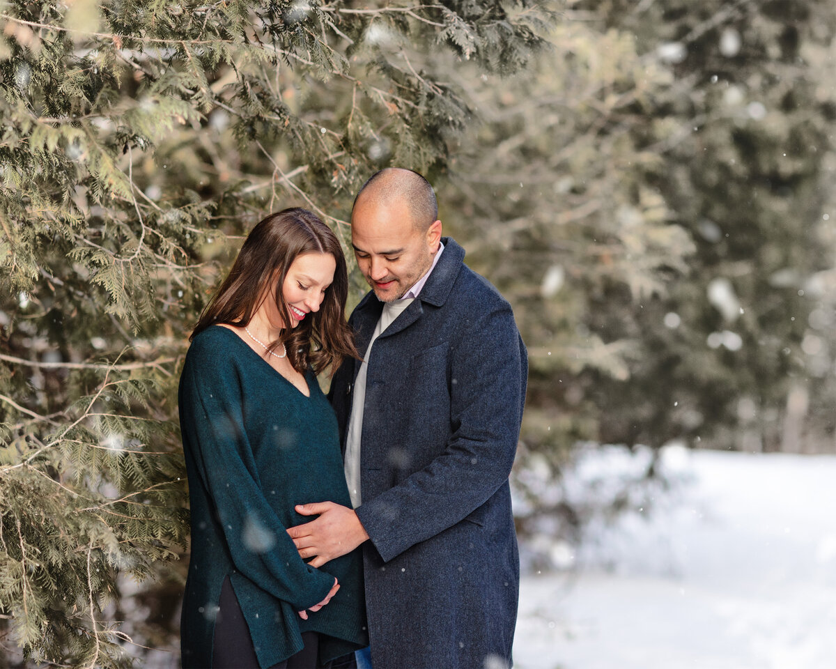 A beautiful outdoor winter maternity portrait photographed after a major snowstorm at Centennial Park in Ayr, Ontario. The expectant couple is surrounded by evergreens as snow gently falls.