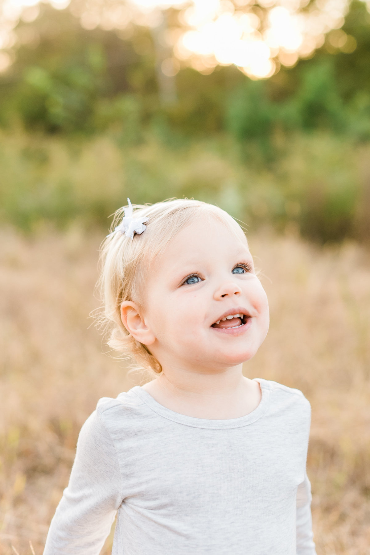 Toddler with blue eyes and a white dress smiles for a portrait during a maternity session in Raleigh. Photographed by Raleigh Maternity Photographer A.J. Dunlap Photography.