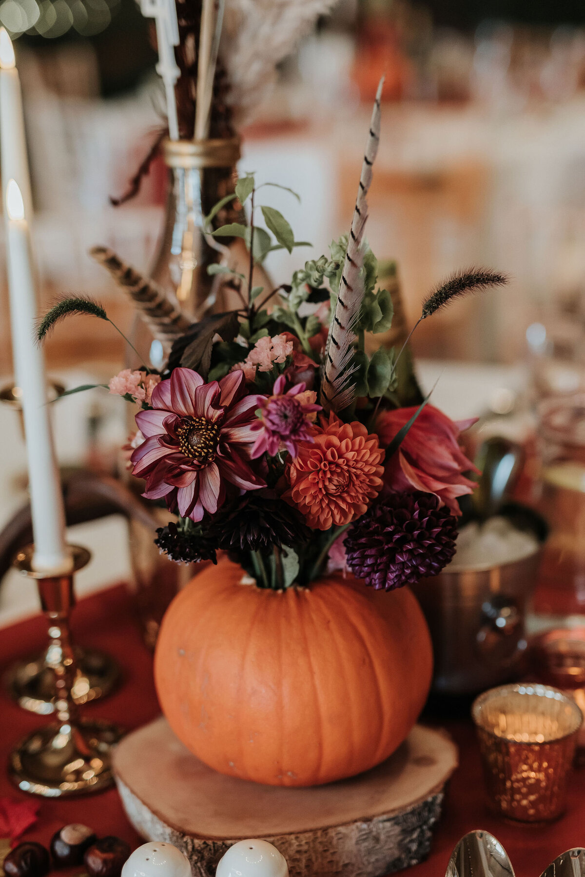 Large orange pumpkin filled with beautiful warm toned wedding flowers and pheasant feather placed on a wooden slice at a rustic Autumn wedding at Montague Farm, Sussex
