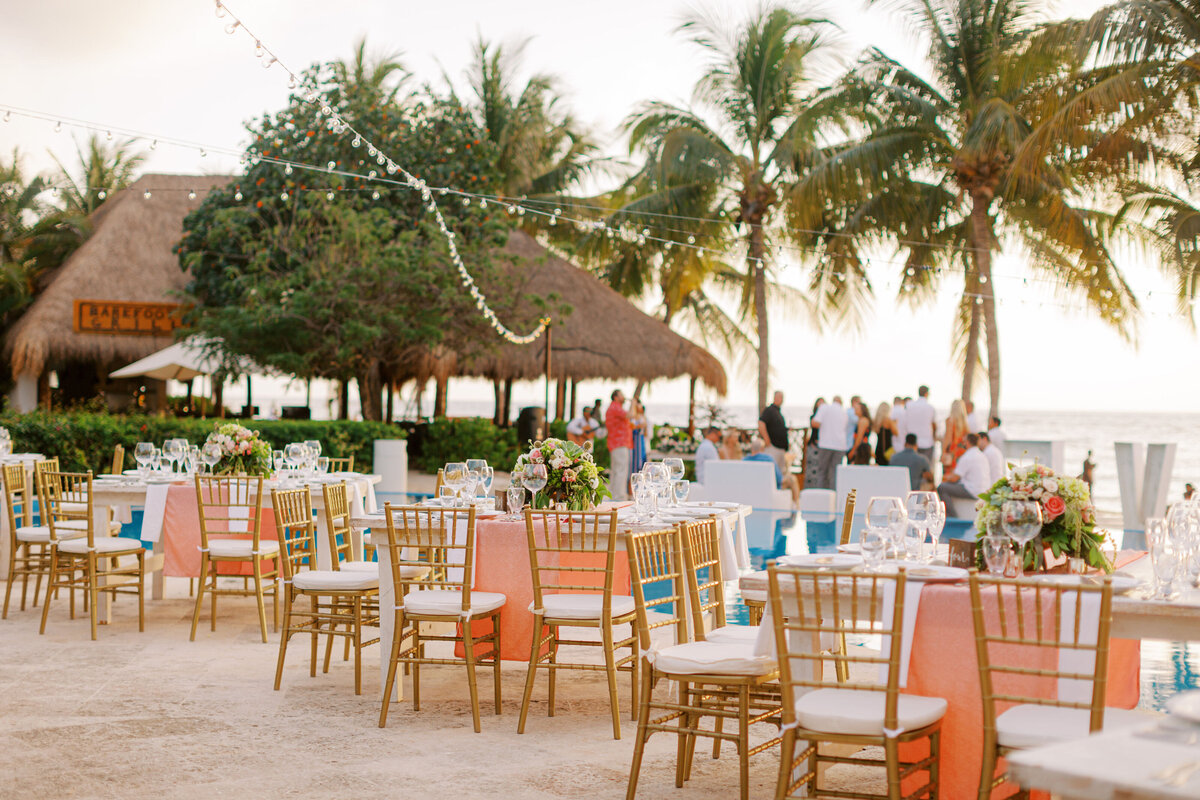Beachfront wedding reception setup with wooden chairs and coral-colored accents under swaying palm trees at sunset.