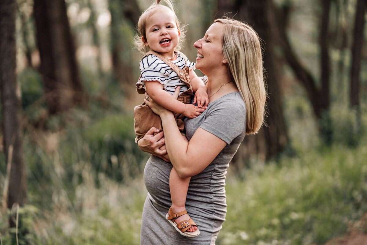 A pregnant mama holds her smiling toddler, straddled above her baby bump during a maternity photo session at the Roberts Bird Sanctuary in Minneapolis.