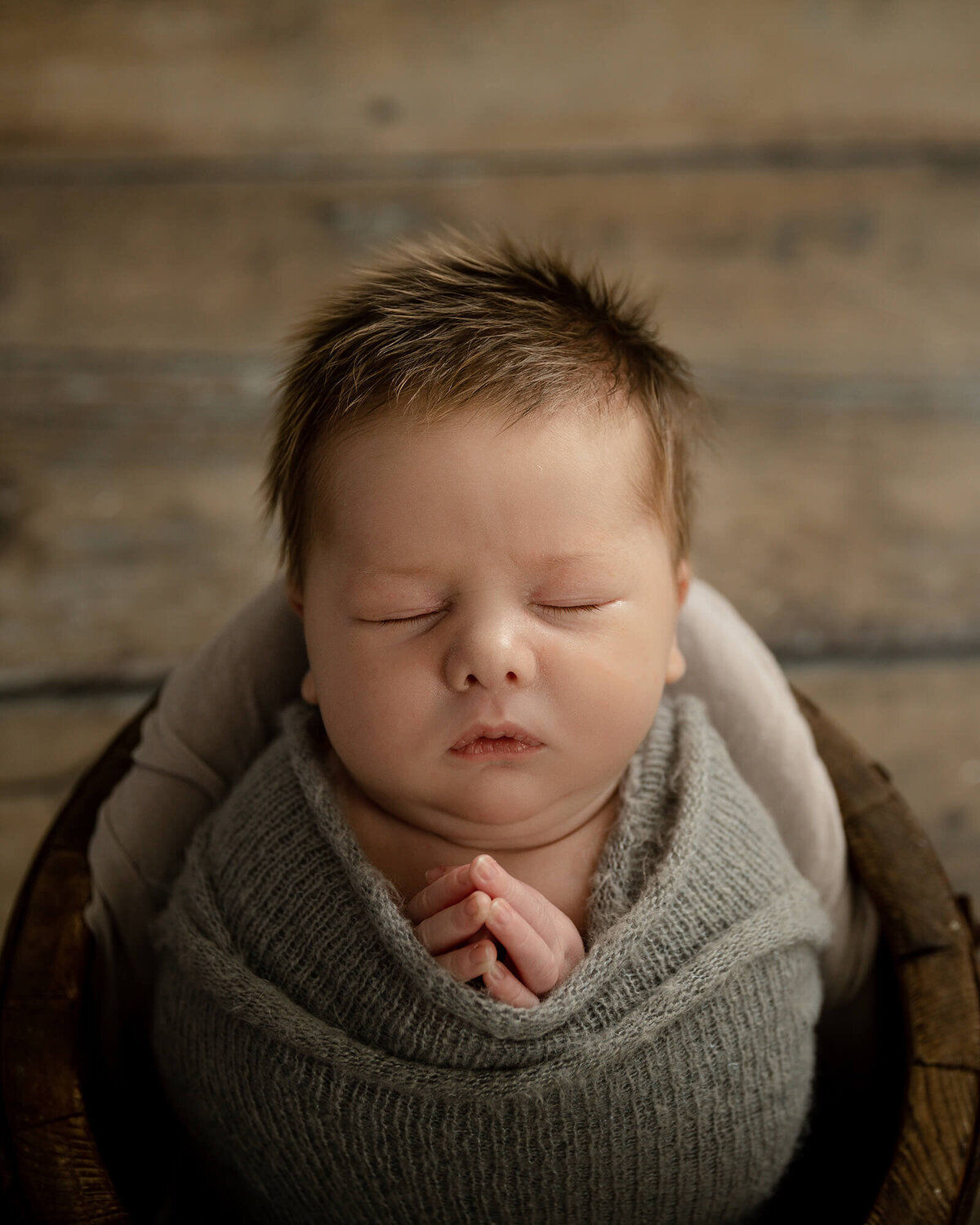 Newborn boy with lots of light brown hair sleeping in a wooden bucket with his hands together like he is praying.