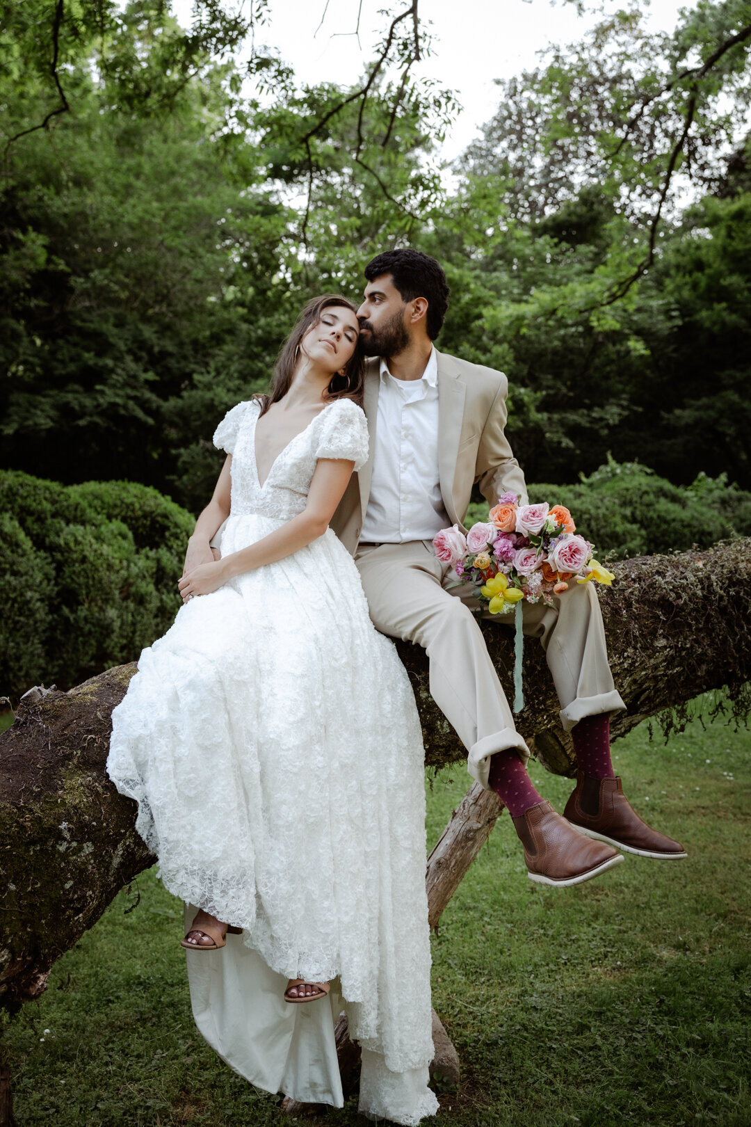 Groom sitting on a tree branch while bride rests her head on his shoulder, holding a vibrant bouquet.