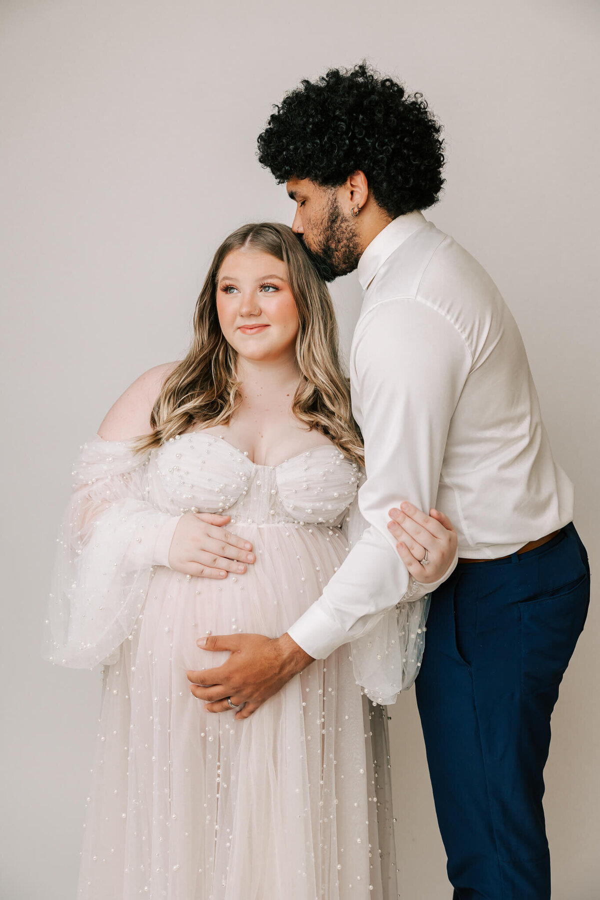 pregnant mom wearing white dress being kissed on the head by her husband