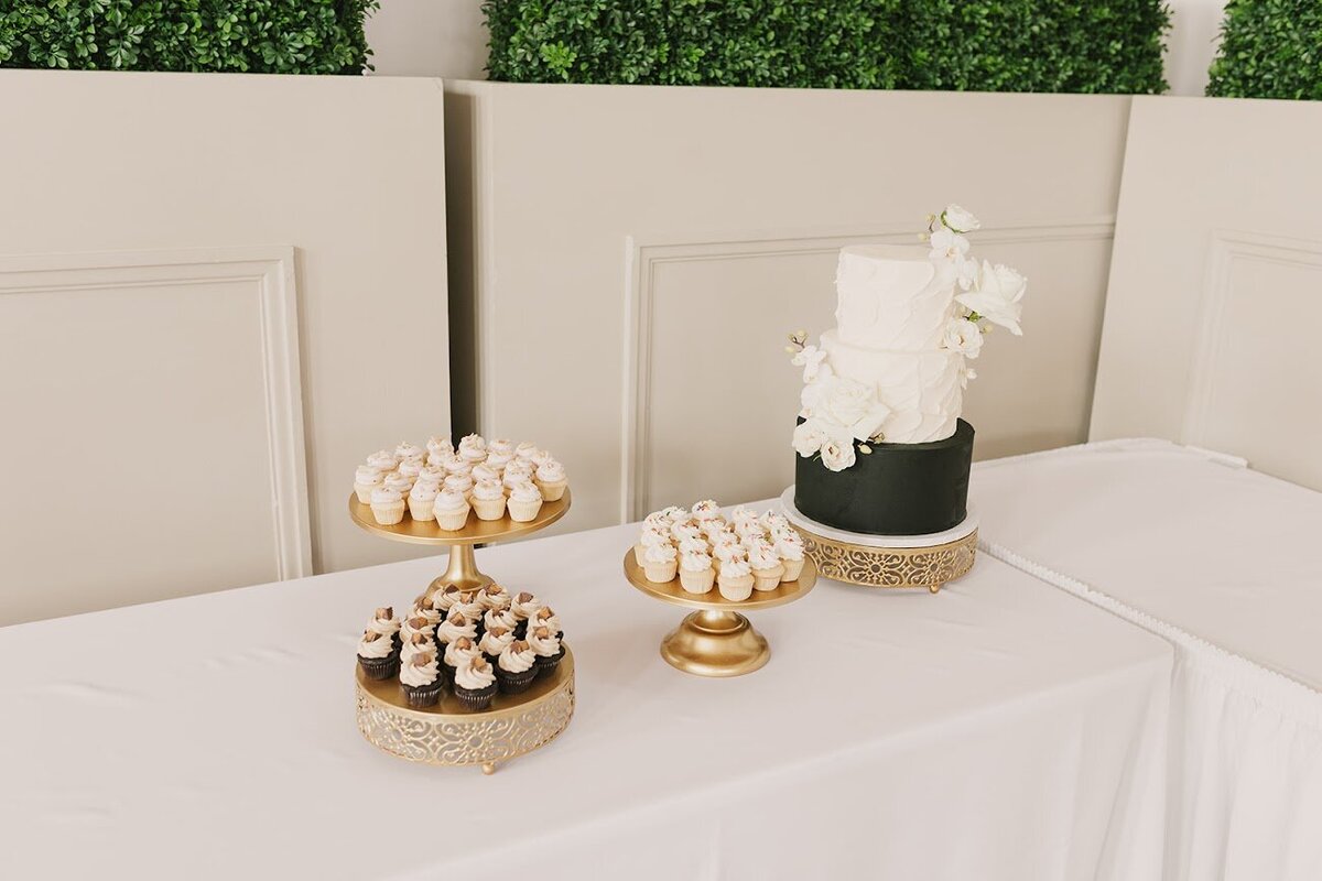 A table with a white tablecloth holds three stands filled with different flavors of mini cupcakes. Near the far corner is a three tier cake with a black bottom tier and two top tiers with white textured frosting and fresh white flower accents. 
