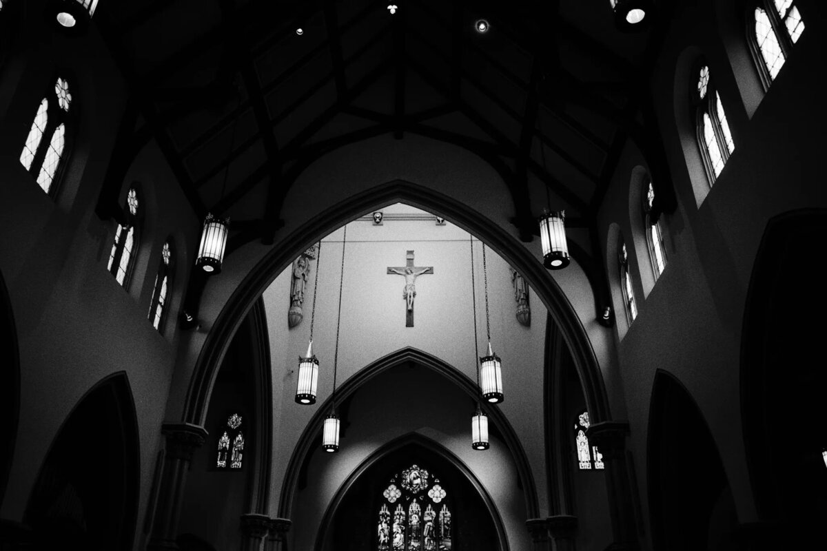 Black and white photo of a church interior by a film photographer NJ, showing high arched ceilings, lanterns, stained glass windows, and a crucifix with Jesus at the center above the altar.
