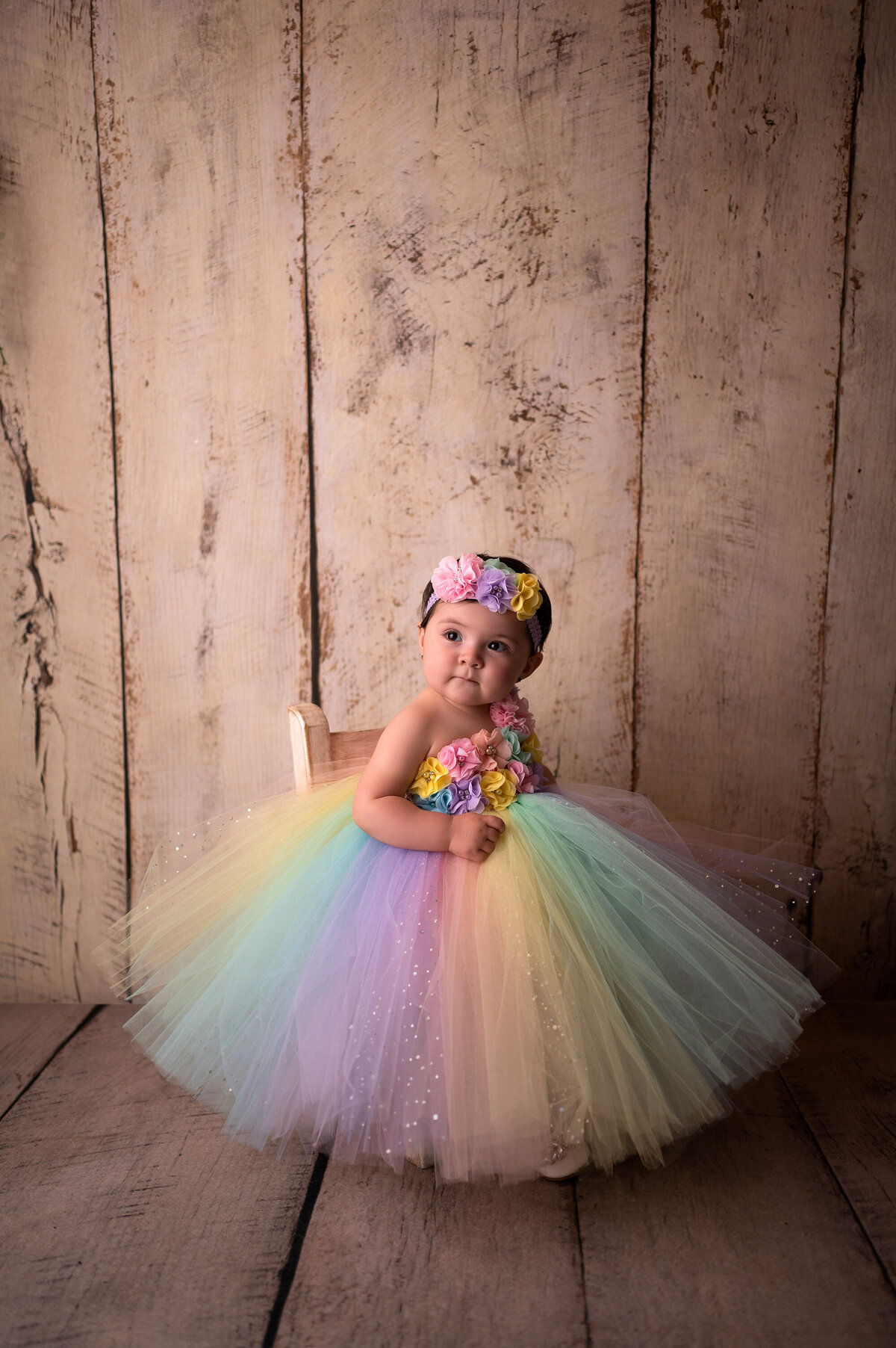 A toddler in a long rainbow tutu sits in front of a paneled wall in our Waukesha, WI portrait studio.