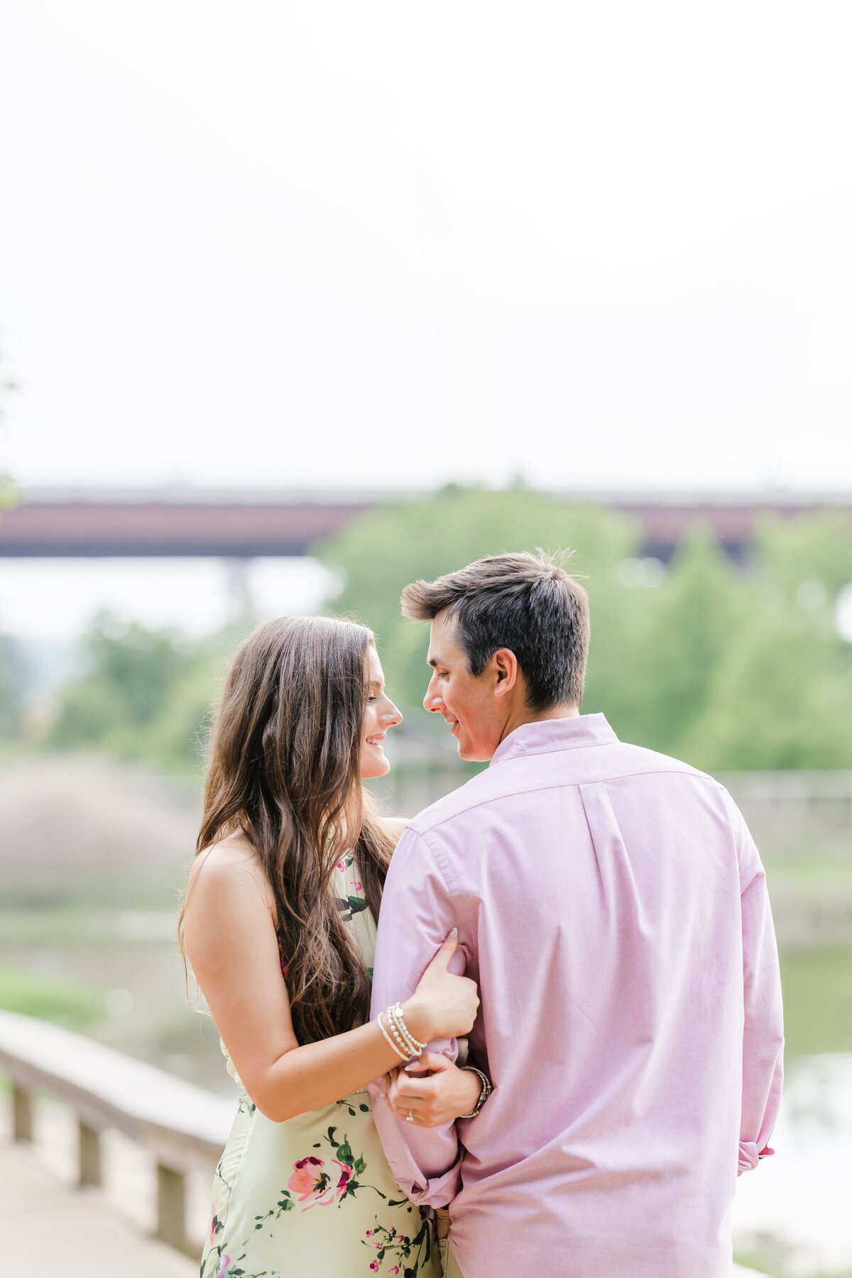A woman in a green floral dress hugs the arm of her fiancee in a pink shirt on a waterfront walkway