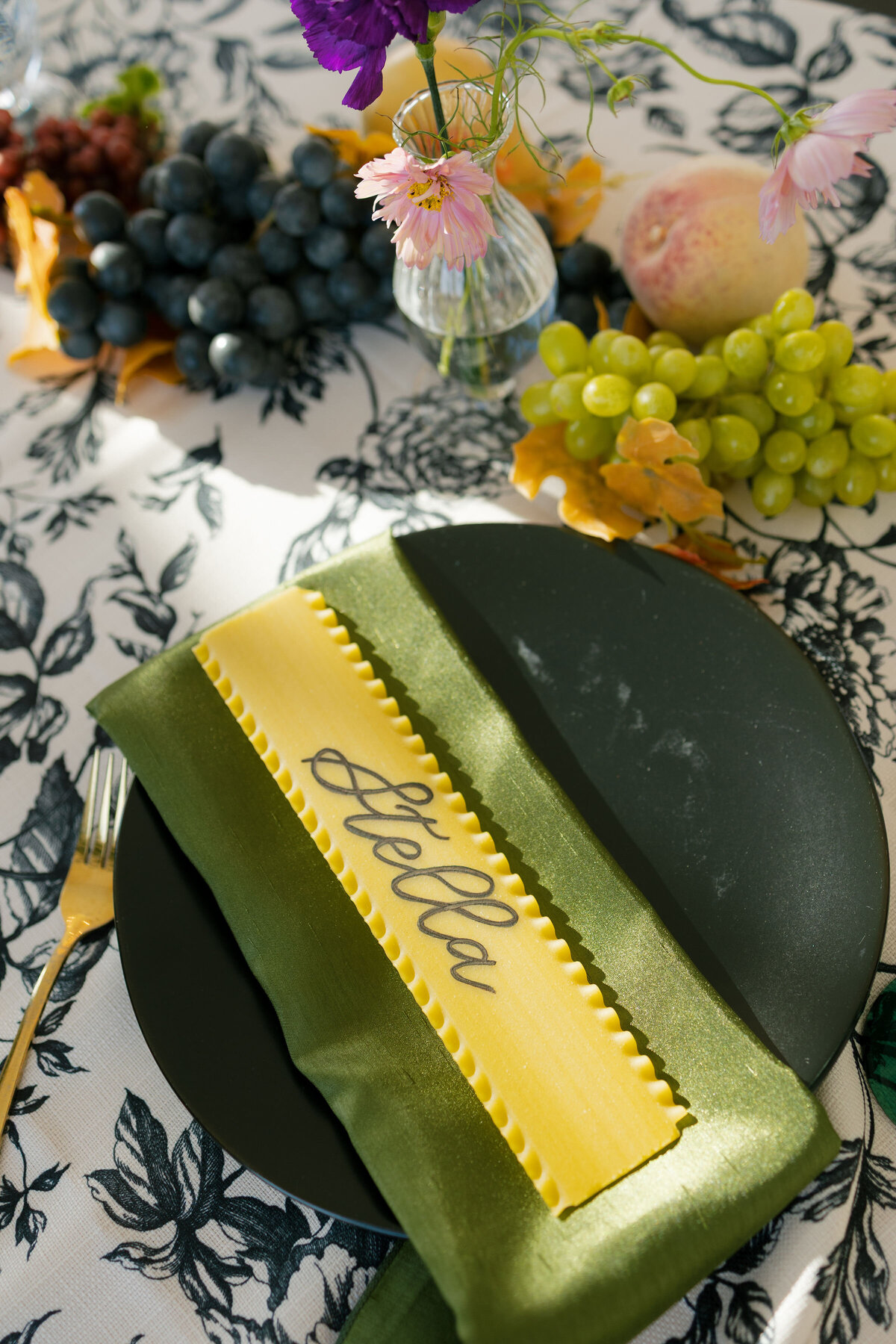 Creative wedding place setting with a handmade yellow pasta name card reading “Stella,” styled atop a green satin napkin on a black plate, surrounded by grapes and delicate bud vase florals.