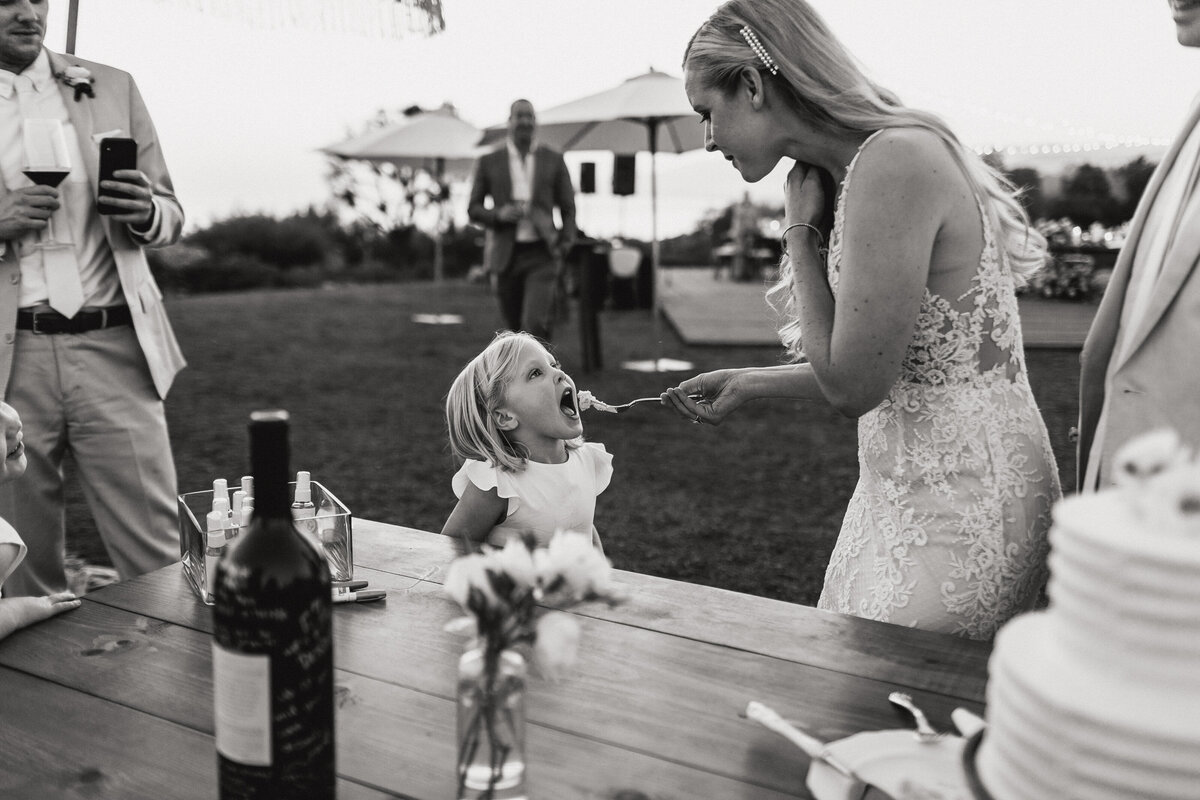 A black and white photograph from Lindsay and Jonsen’s wedding day at Dos Pueblos Orchid Farm in Santa Barbara, California. The bride feeds a forkful of wedding cake to a flower girl who is waiting with her mouth open during their wedding reception. In the foreground is a table with the wedding cake.  Wedding photography by Stacie McChesney/Vitae Weddings.
