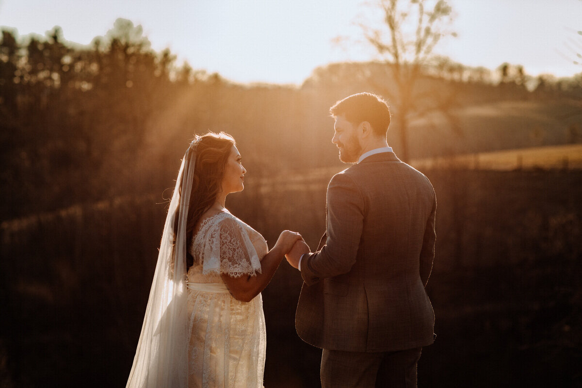 Bride and groom holding hands looking at each other while standing in front of rolling hills with the sunlight coming down on them