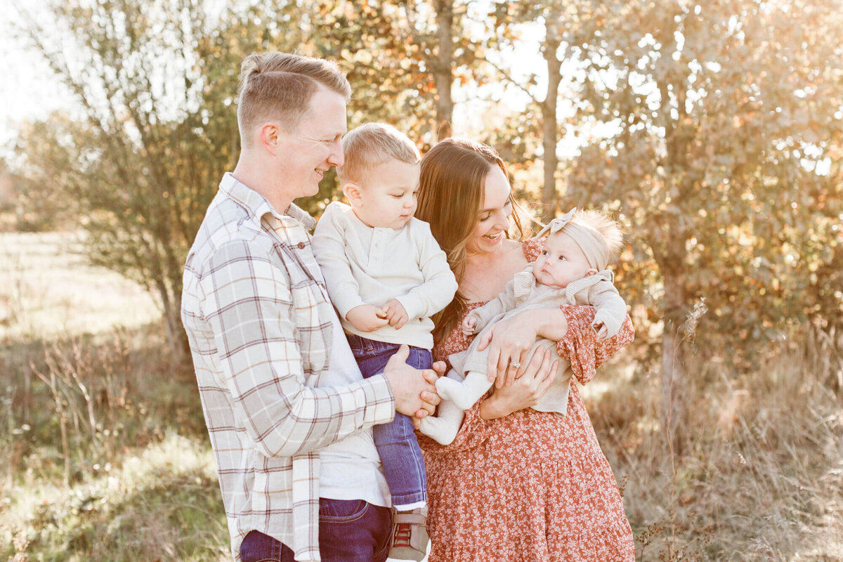 Dad holding toddler and mom holding baby in a golden field at sunset. They are all wearing shades of beige, grey and light pink. 