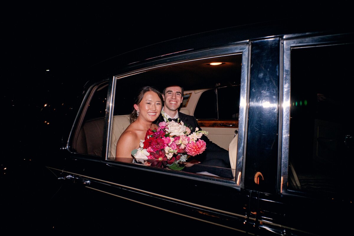 Radiant newlyweds beam from the backseat of a vintage black car, the bride holding a lush bouquet of pink and red blooms. The glow of celebration and romance fills this joyful send-off scene.