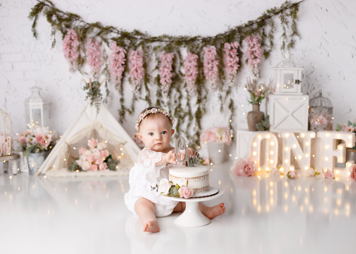 Boho inspired cake smash in West Palm Beach in Boynton Beach newborn photography studio. Baby girl is wearing a white lace romper with a floral crown sitting beside a naked cake decorated with flowers. In the background is a BoHo inspired set up with pink canvas teepee, light up 0NE with a floral banner draped across the back and florals throughout.