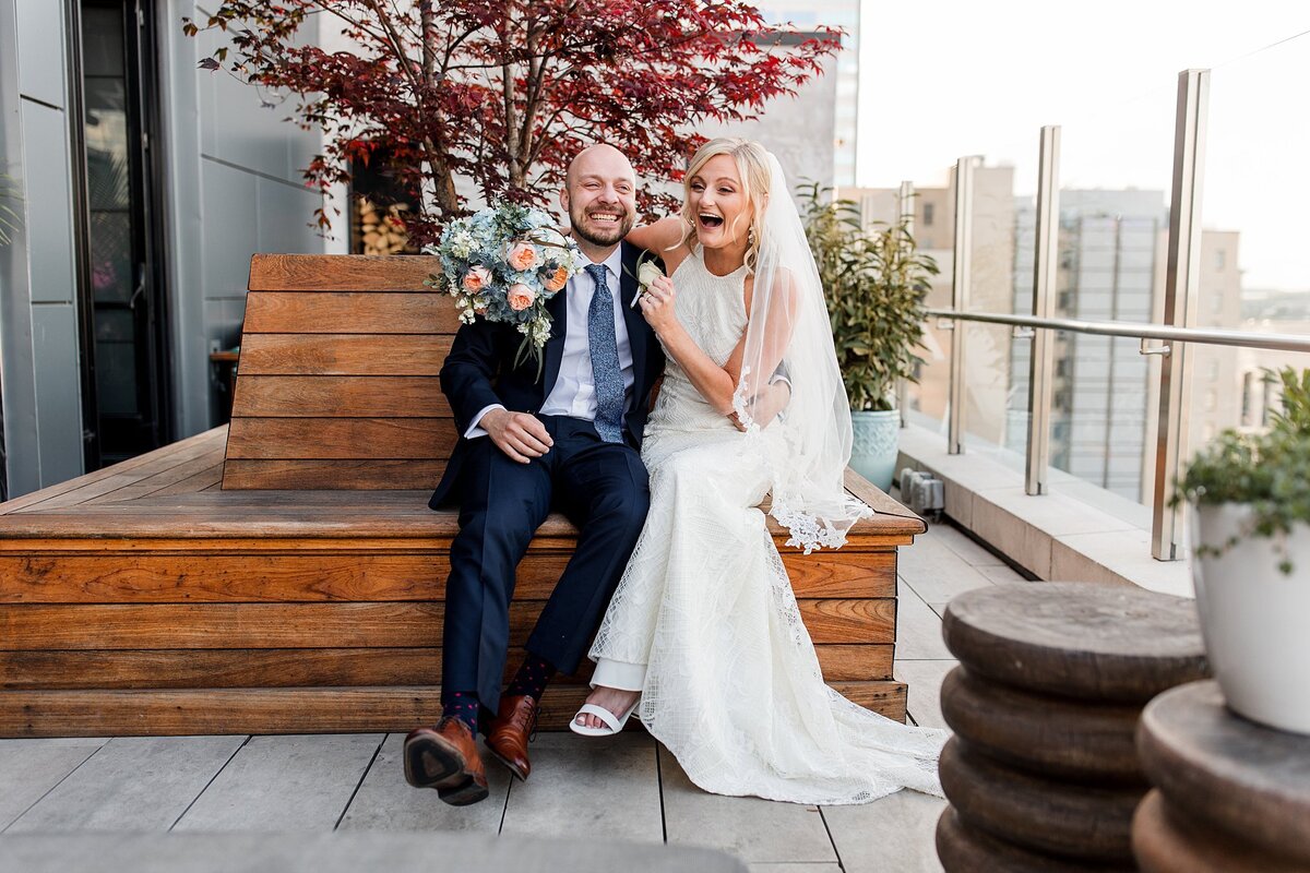 Sitting on a long wooden bench in front of a Japanese maple tree the bride, weaing a fitted halter neck wedding dress and veil shares a laugh with the groom, wearing a navy blue suit and tie. The bride has her arm around the groom's neck and she is holding a bouquet of pink roses, blue hydrangea and ivory roses with greenery at Rare Bird, the rooftop bar at Noelle Hotel in Nashville.