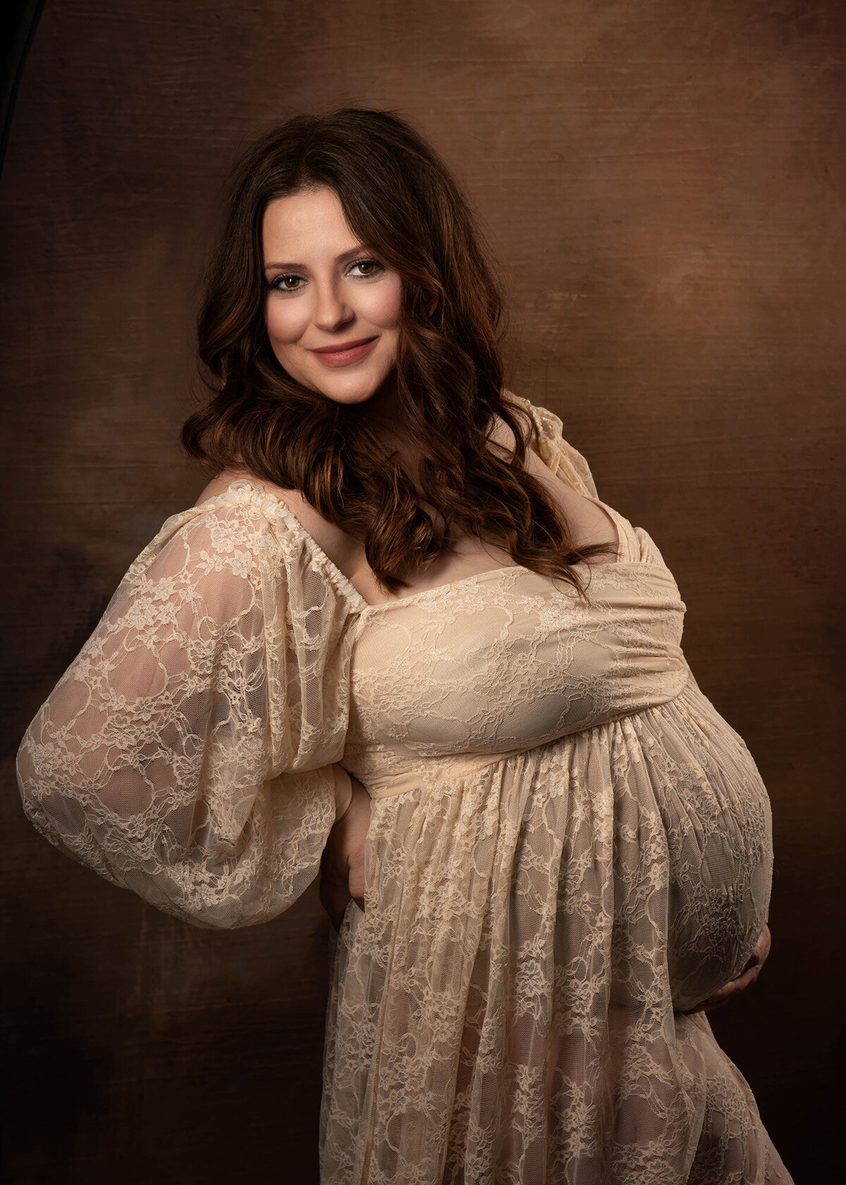 A mother to be stands in a studio in a cream lace maternity gown smiling