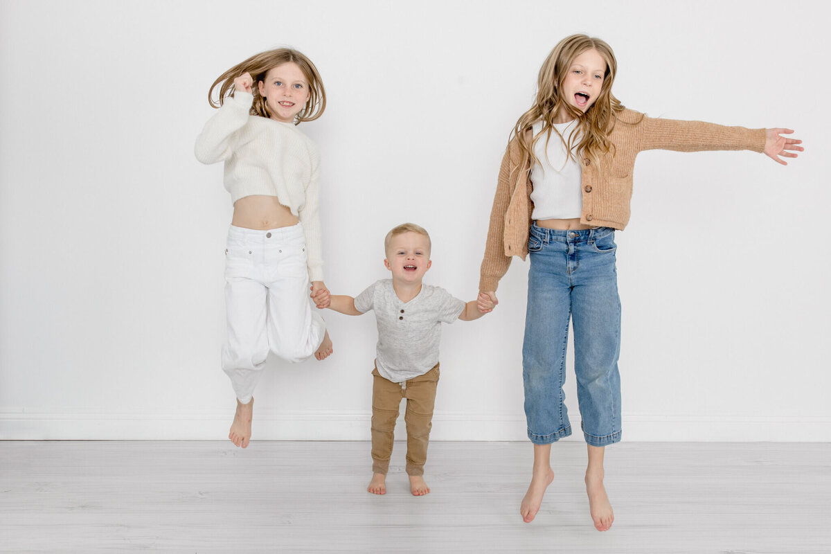 Two young girls with long blonde hair wearing white, blue, and brown, holding their little brother's hands on either side of him, jumping in the air and laughing, looking at the camera.