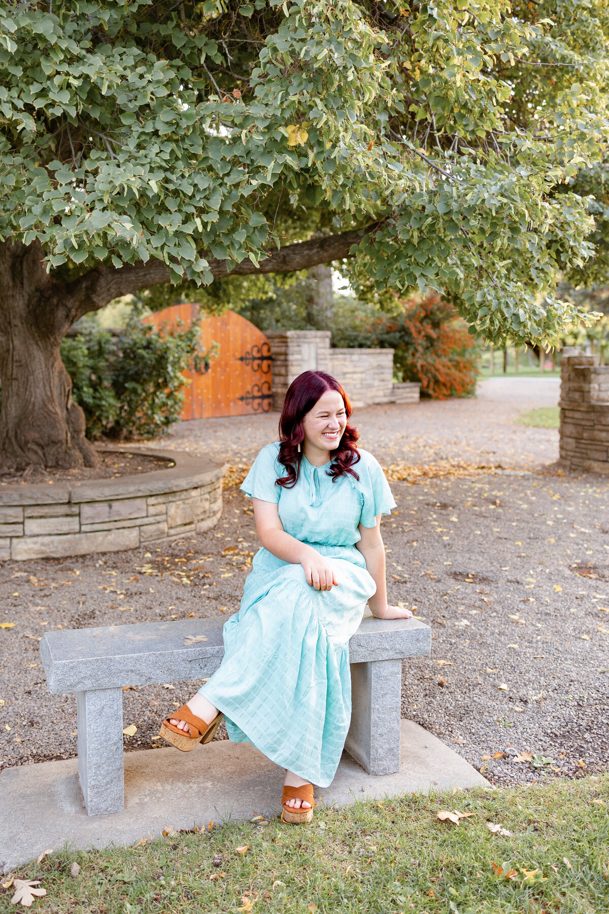 A woman in a light blue dress sits on a stone bench beneath a large tree, enjoying a moment of quiet reflection in a park.
