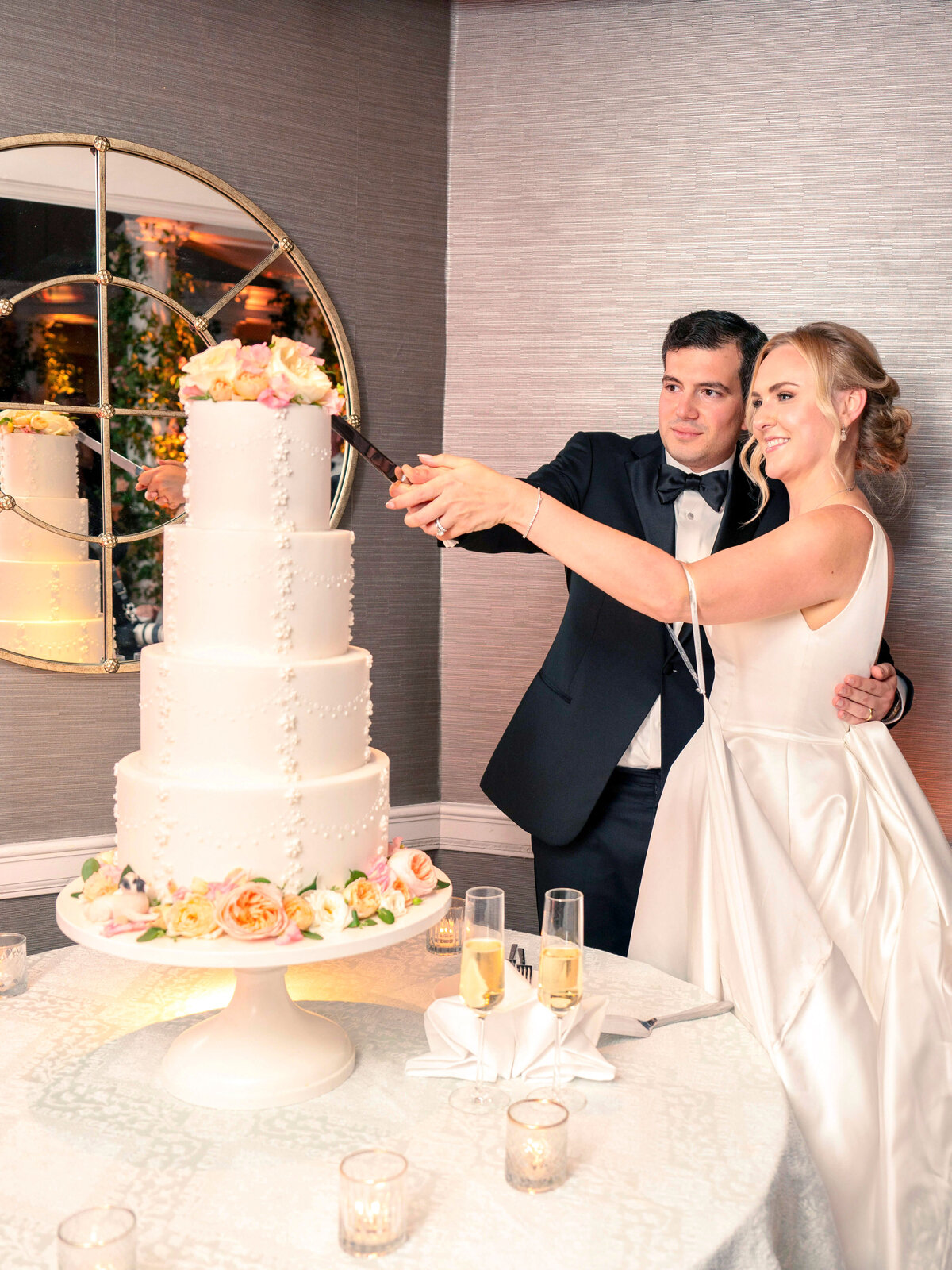 A bride and groom, dressed elegantly, cut a tall, white, multi-tiered wedding cake adorned with flowers on a white stand. Champagne glasses are on the table, and a large round mirror is in the background.