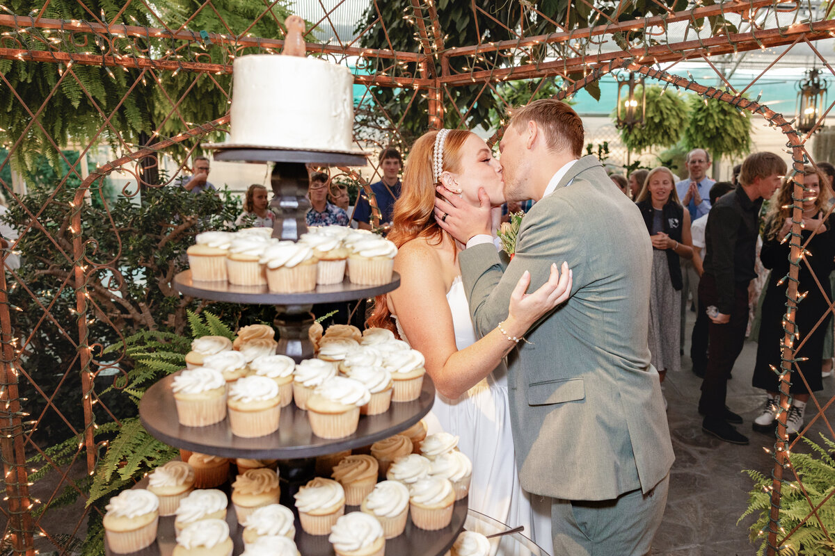 The bride and groom share a romantic kiss in front of their wedding cake, with guests watching in celebration