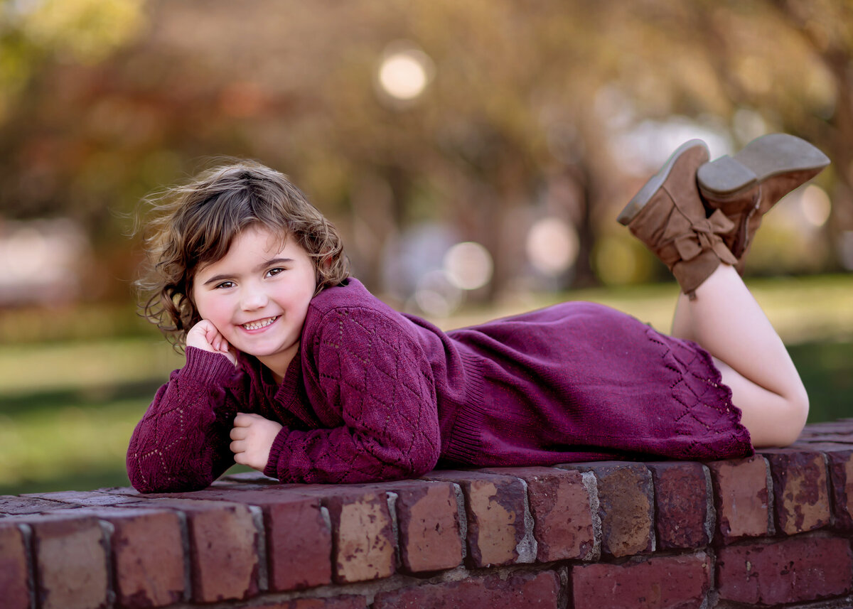 Child in a purple sweater dress lying on a brick wall with autumn foliage in the background.