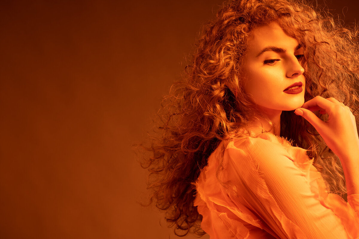 Portrait of a woman with voluminous curly hair, bathed in warm, golden-orange lighting.