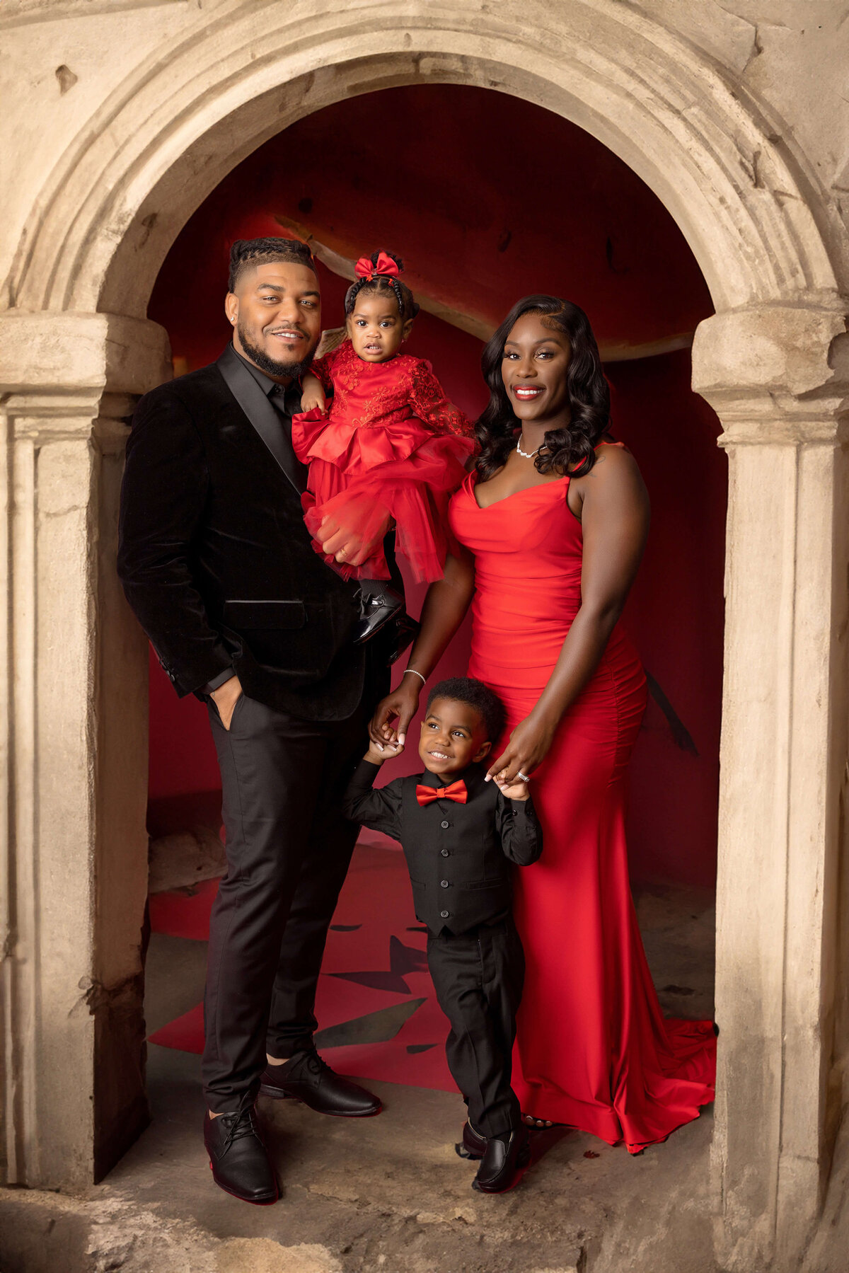 A family of four poses inside a stone archway, all dressed in red and black formal attire. The father holds a young girl in a red dress, while the mother in a red gown stands next to a small boy in a black suit with a red bow tie—captured perfectly by Jacksonville family photography experts.