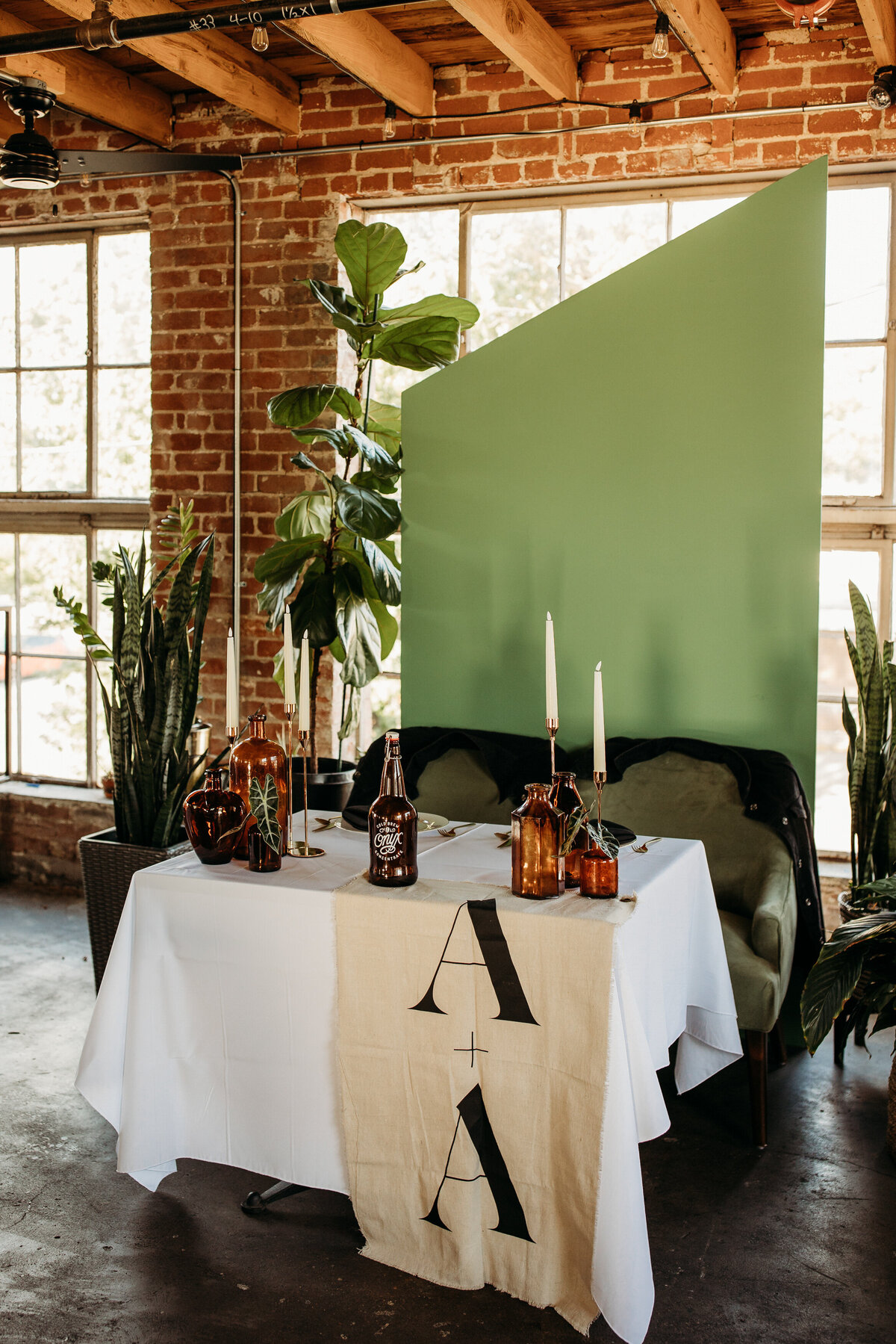 Rustic wedding table setup featuring a green backdrop, brown glass bottles with candles, and a tablecloth with bold typography