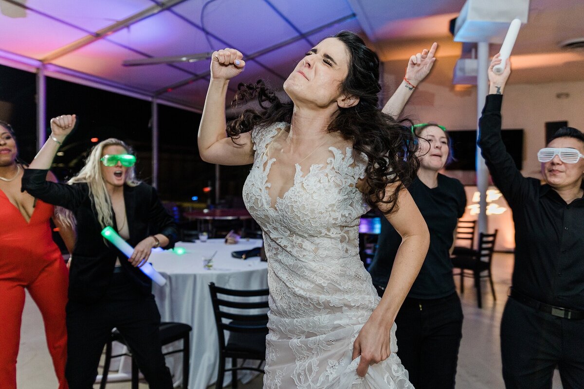 A bride enthusiastically dancing during her wedding reception in Dallas, Texas. The bride is wearing a short sleeve, intricate, white dress, and some of her wedding guests can be seen dancing behind her and cheering her on.