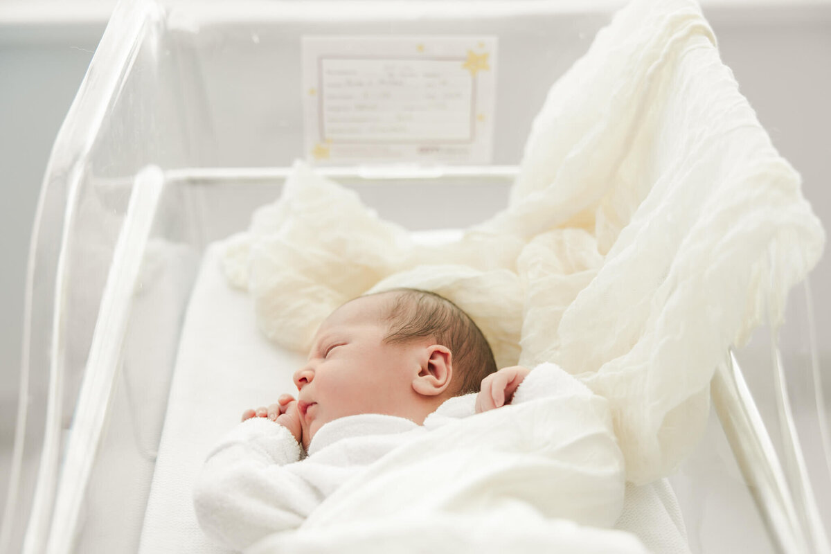 Cute photo of a newborn baby lying in his hospital crib, swaddled by white and cream blankets.
