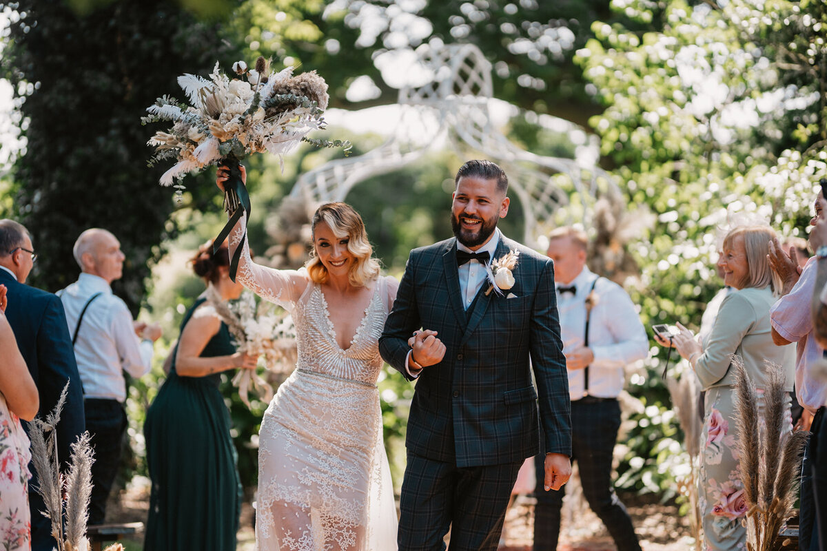  A joyful bride and groom walk hand-in-hand down the aisle after their outdoor wedding ceremony. The bride, wearing a sparkling white gown, holds a bouquet triumphantly in the air and smiles at the camera, while the groom, in a checked suit with a boutonnière, beams with happiness. Guests in the background clap and celebrate amidst lush greenery and sunlight.