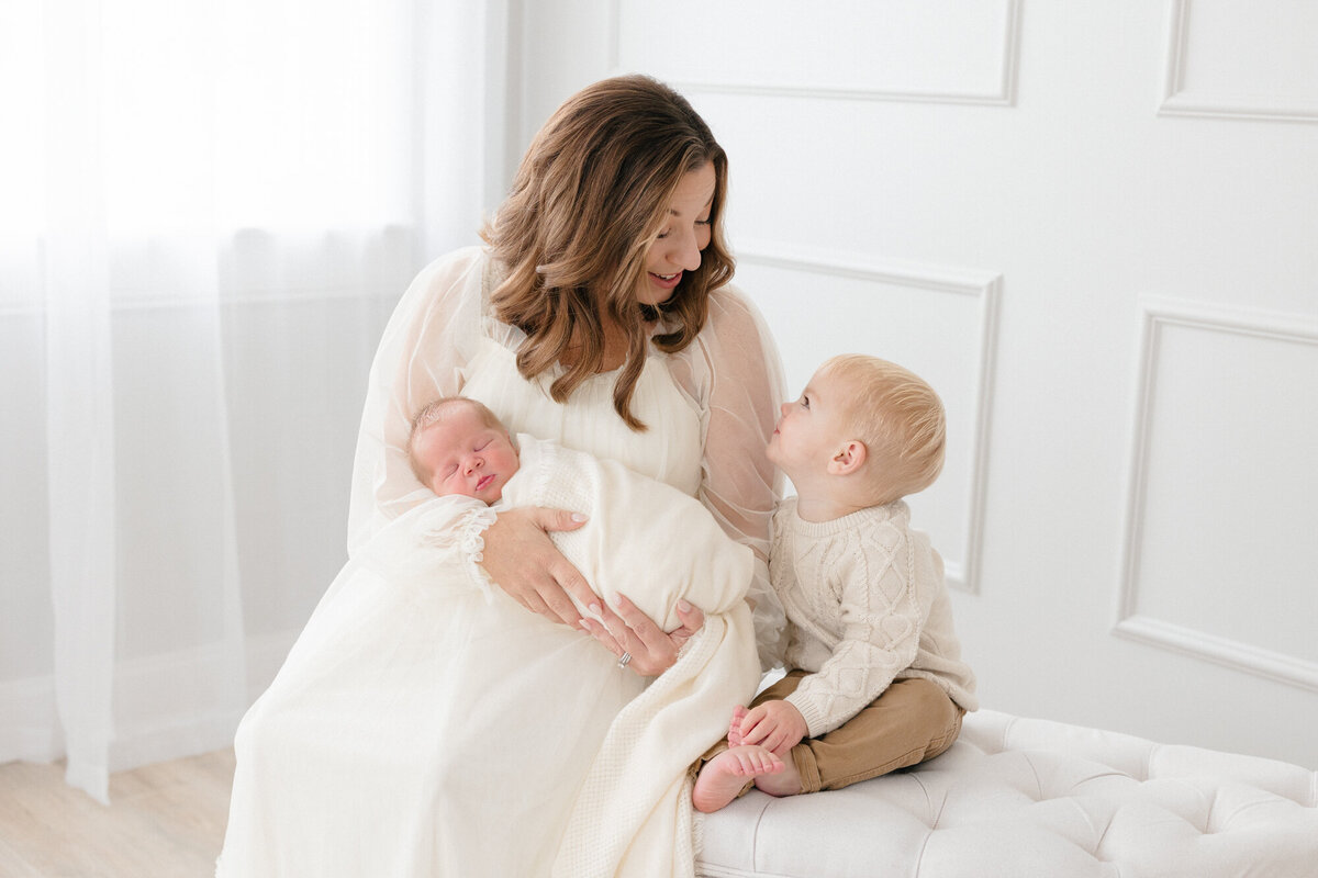 Mom holding her newborn baby boy and smiling at her older child in a louisville newborn photography studio