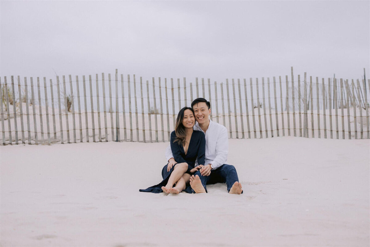 The engaged couple is happily sitting on the smooth, white sands of Fire Island Beach, NY. Engagement Image by Jenny Fu Studio