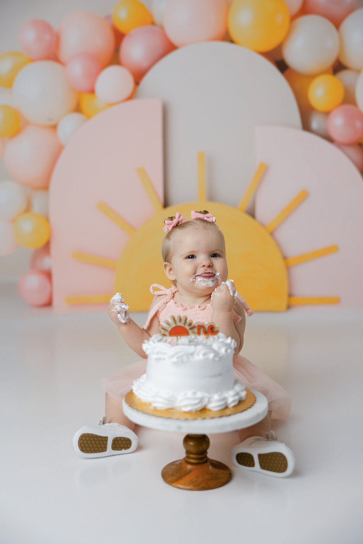 Baby in peach “one” dress enjoying cake smash with sunrise backdrop and pastel balloons. Photograph by Yucaipa cake smash photographer Kaitlyn Dawn Photography.