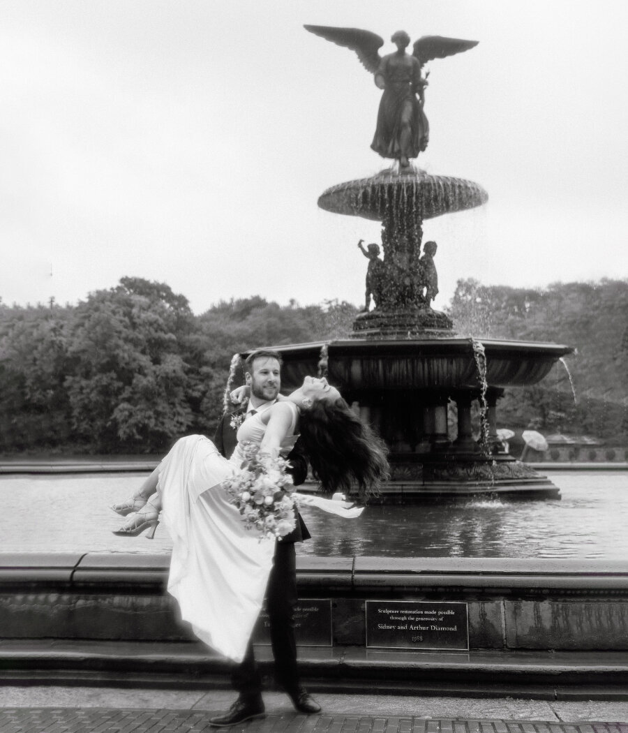 Groom-to-be lifting his fiancée in front of Bethesda Fountain in Central Park after surprise proposal, captured in joyful black and white